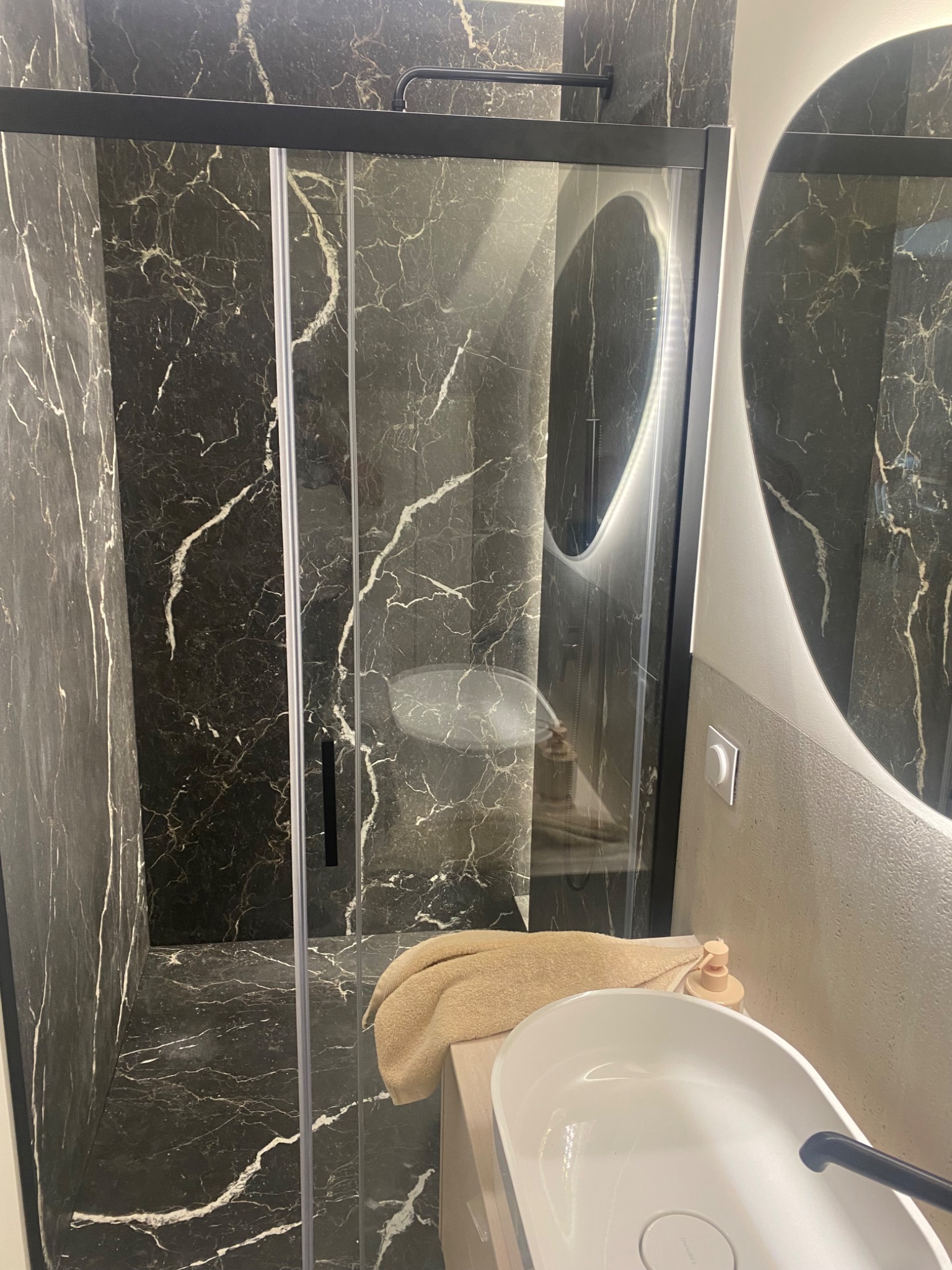Nowoczesna łazienka z czarnymi płytkami imitującymi marmur, kabiną prysznicową typu walk-in z czarnymi profilami i okrągłym lustrem z podświetleniem LED.
