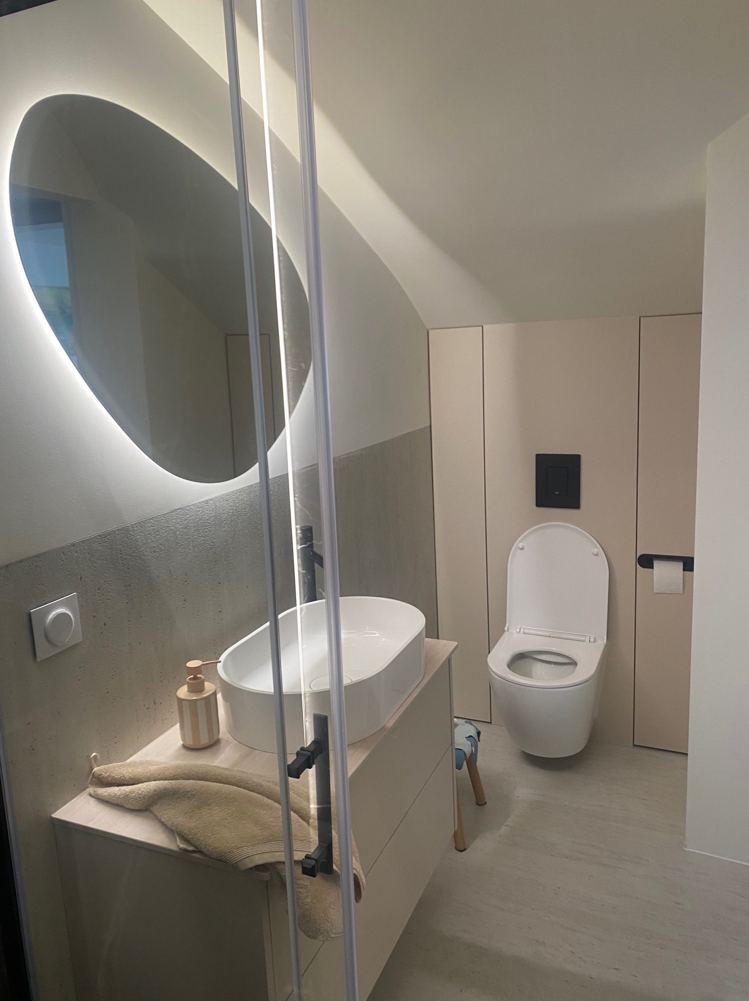 Nowoczesna łazienka z owalnym lustrem podświetlanym LED, umywalką nablatową i toaletą w zabudowie. Minimalistyczny design z elementami betonu architektonicznego i jasnych odcieni.