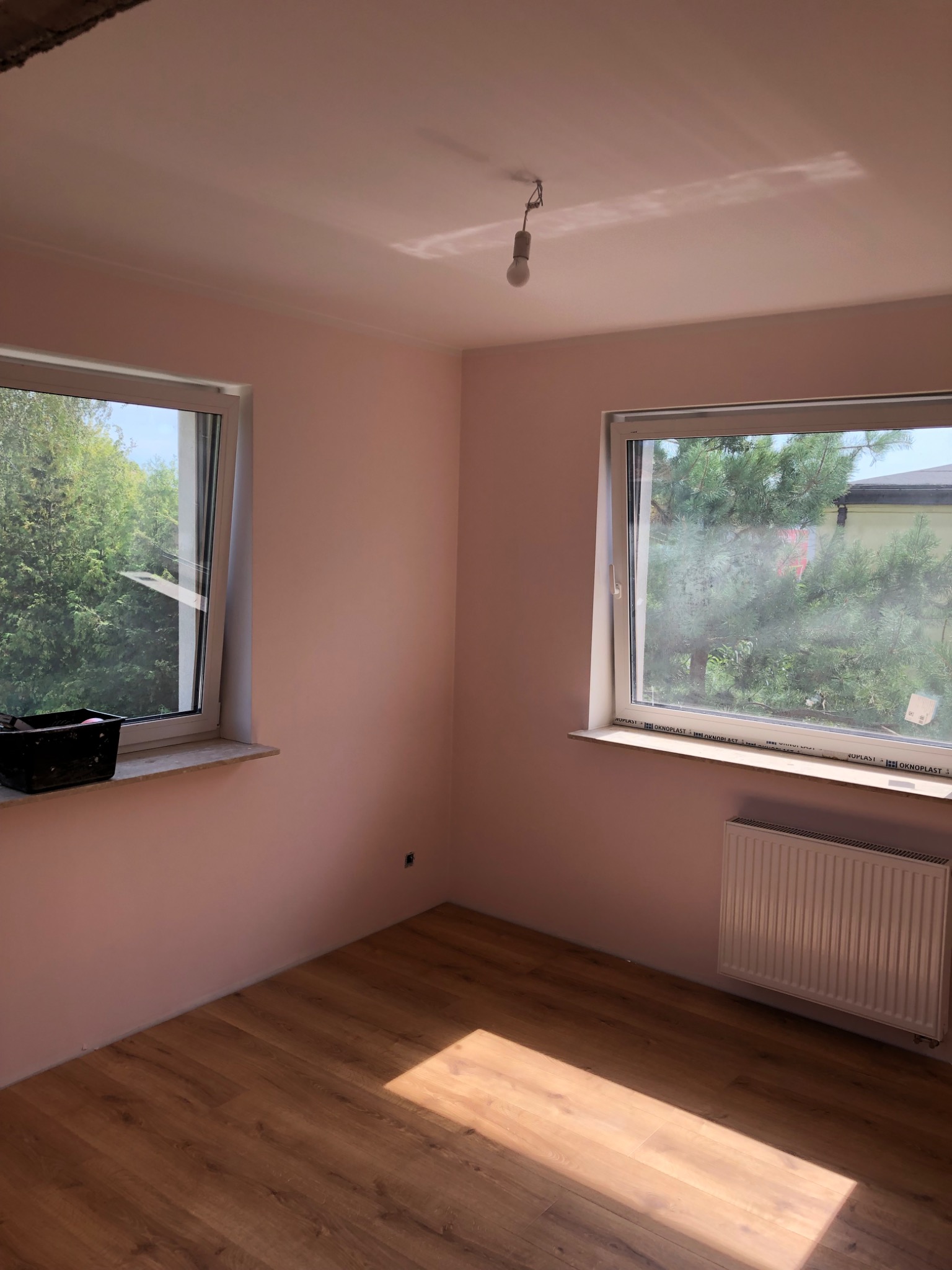 Pokój po malowaniu ścian na różowo, z drewnianą podłogą i dwoma oknami. Na parapecie stoi czarny pojemnik malarski. Widoczny grzejnik i wisząca żarówka.