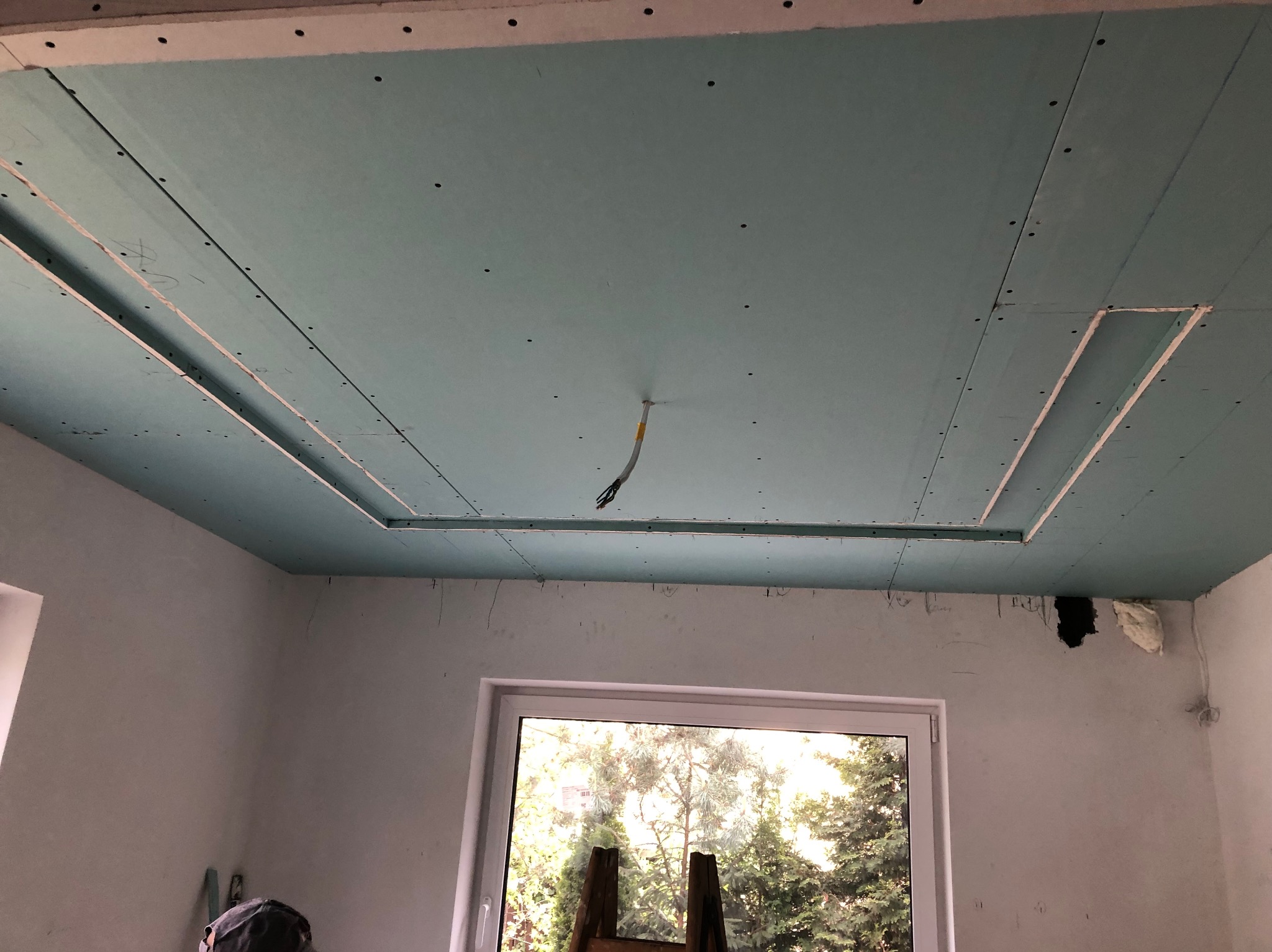 Niebieski sufit z płyt kartonowo-gipsowych w trakcie montażu, widoczne mocowania i przewody. W tle okno z widokiem na zieleń.