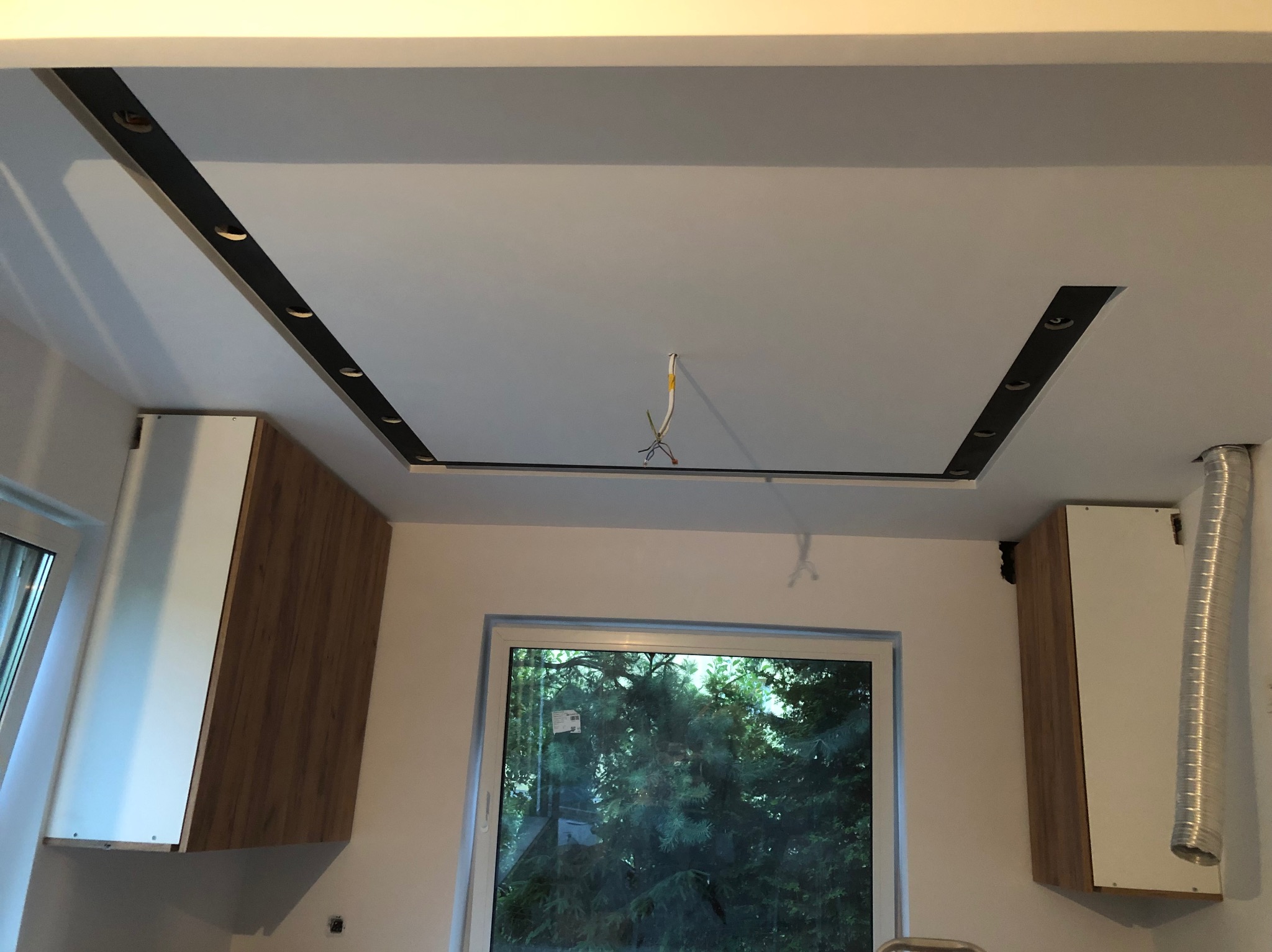 Nowoczesny sufit podwieszany z czarnymi listwami i otworami na oświetlenie punktowe w kuchni, widoczne szafki i okno z widokiem na zieleń.