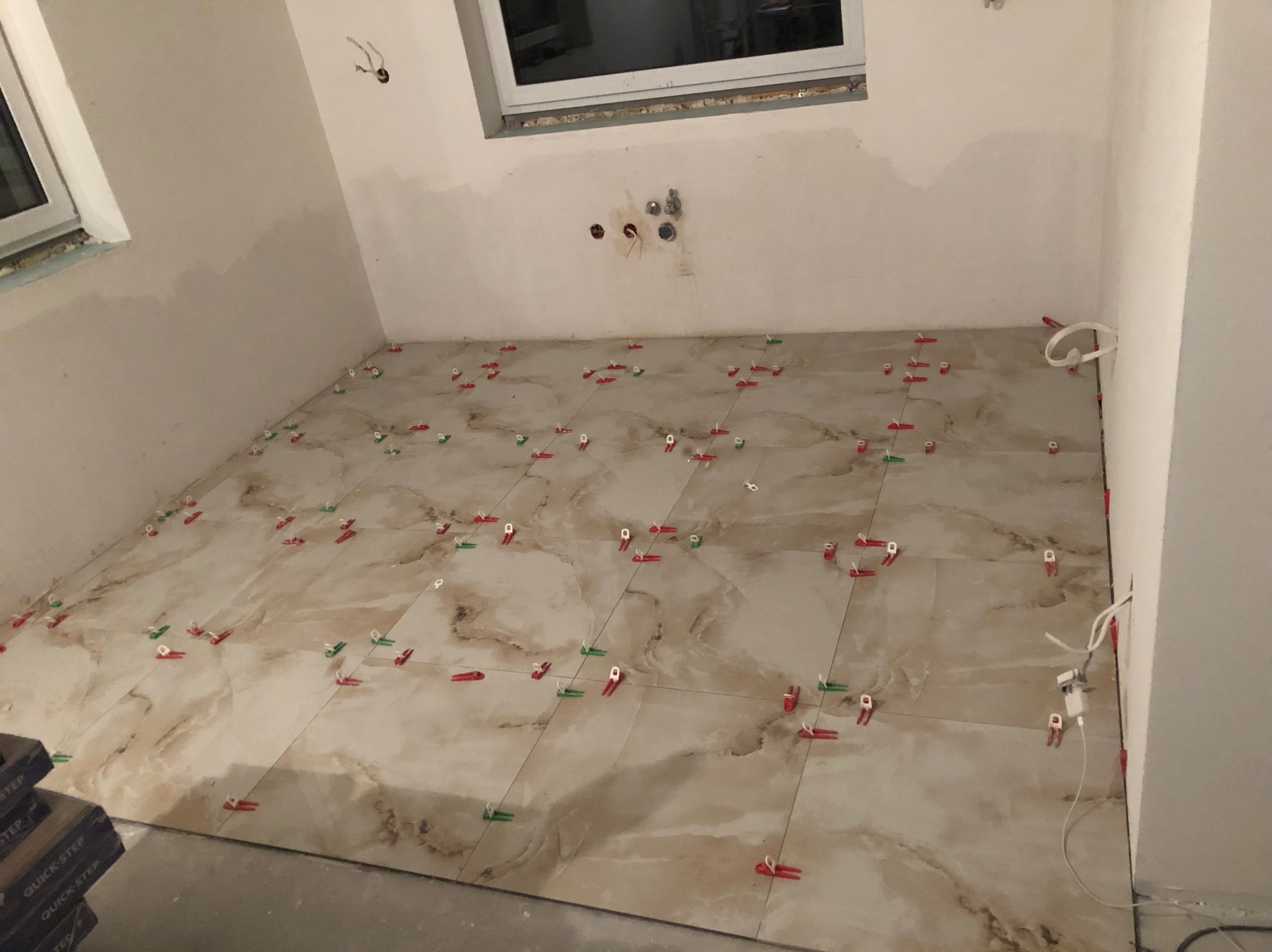 Układanie płytek podłogowych w jasnym odcieniu z użyciem klipsów poziomujących w trakcie remontu łazienki. Widoczne okno i instalacja elektryczna.