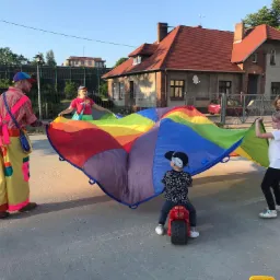 Animatorzy w kolorowych strojach rozciągają tęczową płachtę nad dziećmi, w tle budynek z czerwonym dachem.