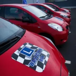 Rząd czerwonych samochodów z naklejkami 'L' i logotypem 'WASZCZYŃ OSK' na masce, ustawionych na parkingu przed budynkiem.