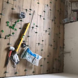 nie podano - Montaż drewnopodobnych płytek na ścianie z użyciem systemu poziomowania; widoczne narzędzia: poziomica, młotek, pojemnik z klinami, rękawice robocze oraz worek z systemem poziomowania.