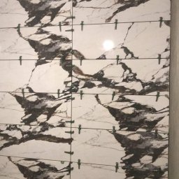 nie podano - Montaż płytek imitujących marmur na ścianie, widoczne zielone krzyżyki dystansowe, surowy stan wykończenia, szara ściana w tle.