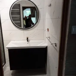 Wykończona łazienka z umywalką na szafce w kolorze czarnym, okrągłym lustrem z widocznym grzejnikiem drabinkowym w odbiciu, białe płytki imitujące marmur na ścianach, oraz zamontowane wieszaki...