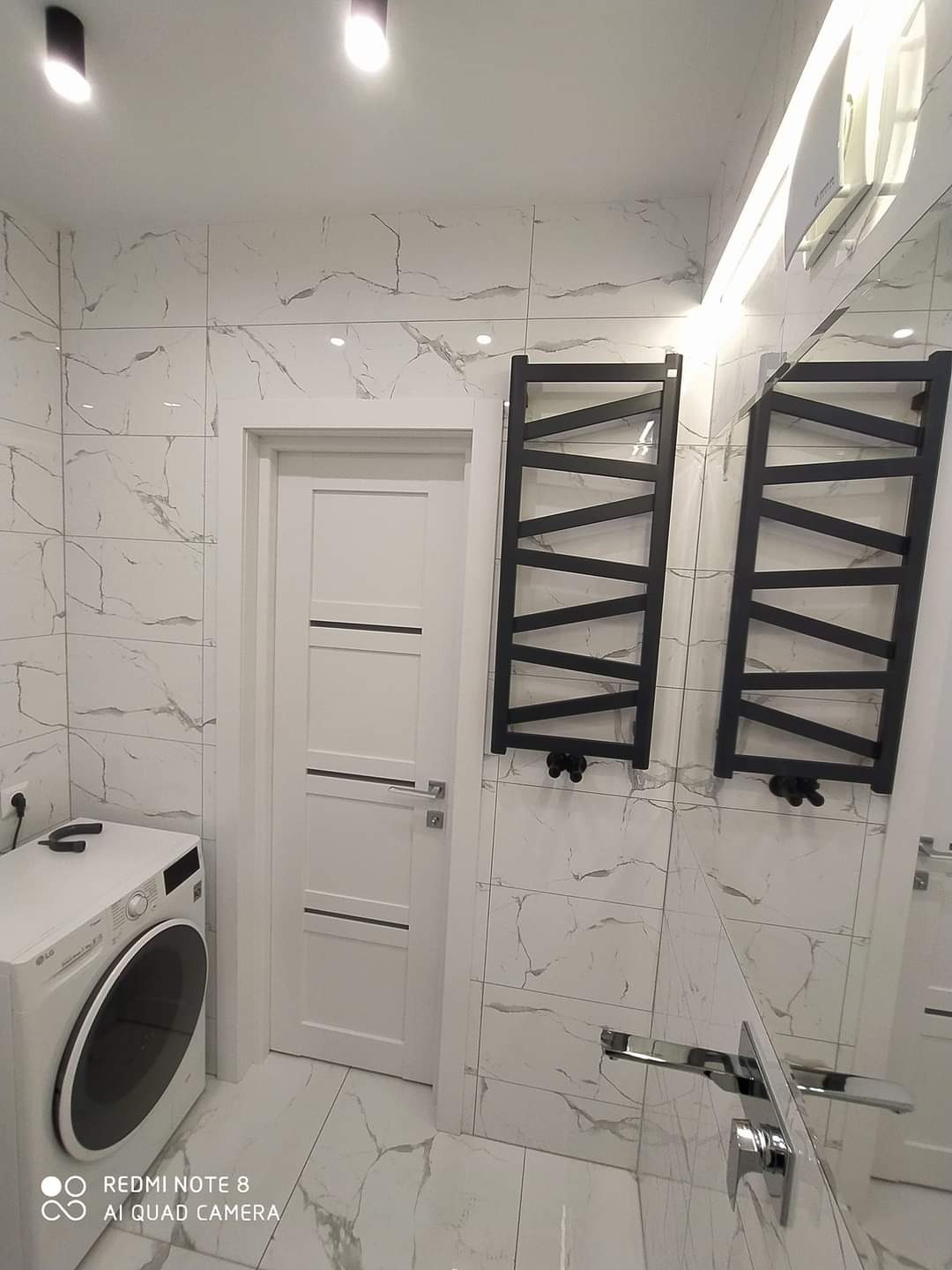 Wykończona łazienka z biało-szarymi płytkami imitującymi marmur, białą pralką LG, białymi drzwiami wewnętrznymi z poziomymi frezami i dwoma czarnymi grzejnikami drabinkowymi na ścianie.