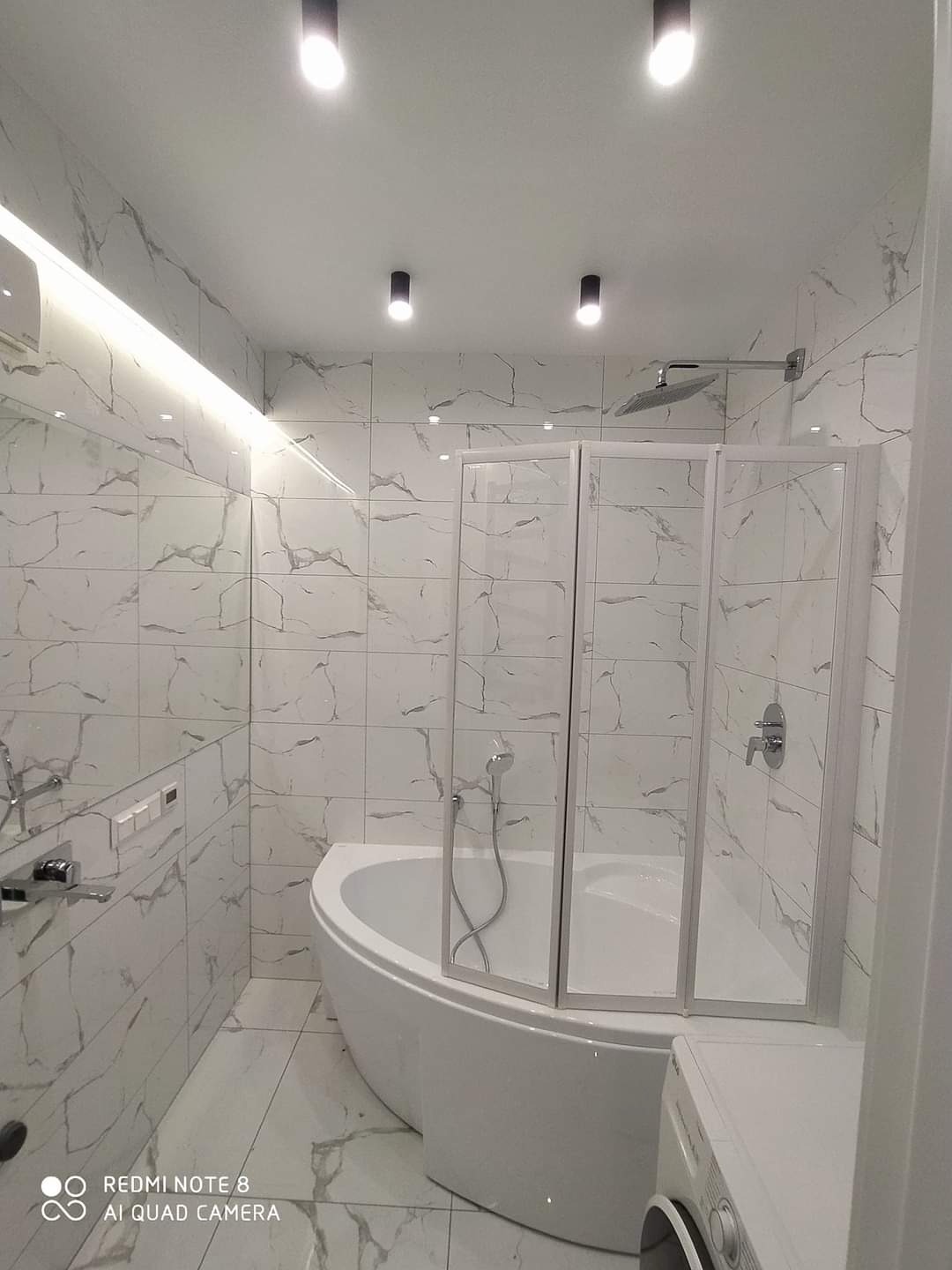 Nowoczesna łazienka z narożną wanną obudowaną szybami, wykończona białymi płytkami imitującymi marmur, oświetlona punktowymi lampami sufitowymi i listwą LED nad lustrem.