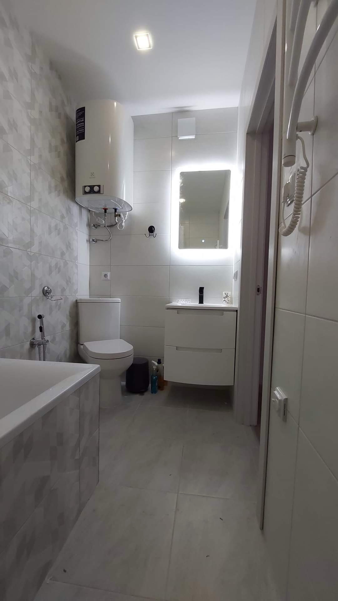 Nowoczesna łazienka z białymi płytkami o wzorze geometrycznym na ścianie, podświetlanym lustrem nad umywalką z szafką, bojlerem elektrycznym i grzejnikiem drabinkowym.