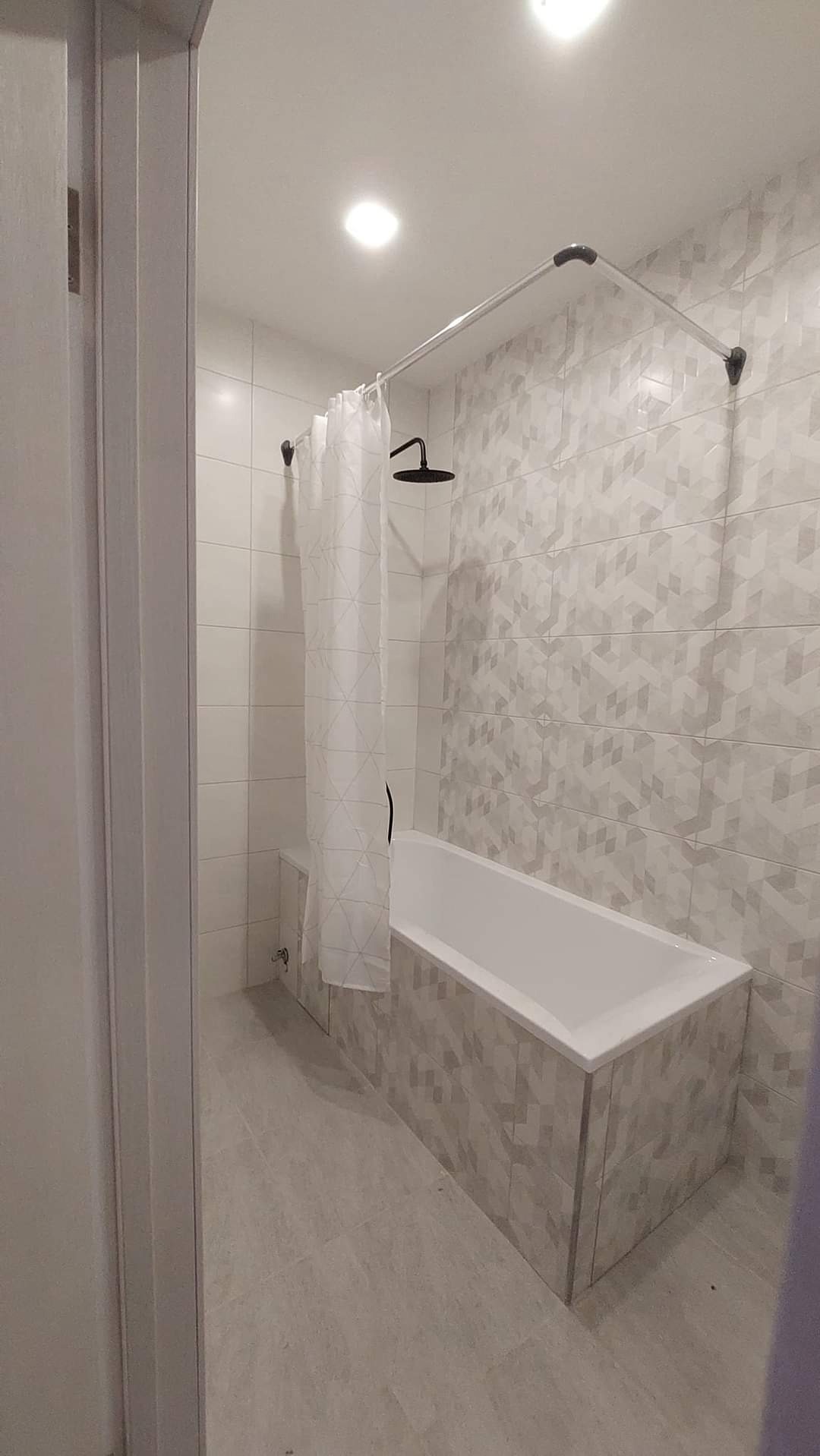 Nowoczesna łazienka z wanną obudowaną płytkami w geometryczny wzór, biała zasłona prysznicowa z delikatnym nadrukiem, czarna deszczownica i jasne płytki na ścianach.