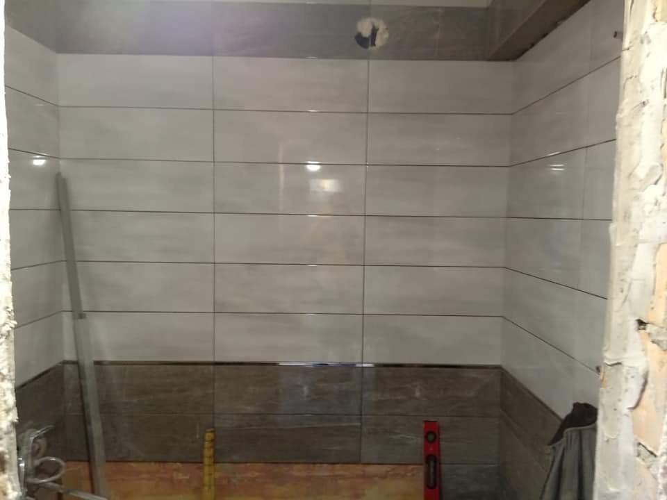 Wnętrze łazienki w trakcie remontu: ułożone jasnoszare płytki ścienne z ciemniejszym pasem dolnym, widoczna poziomica, fragment ściany w stanie surowym.