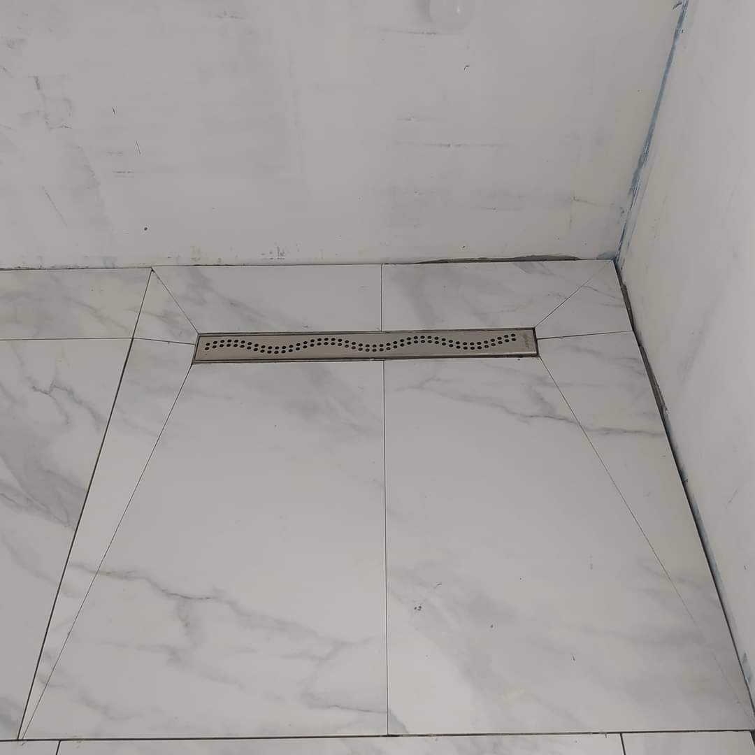 Białe płytki imitujące marmur ułożone ze spadkiem w kierunku liniowego odpływu w kabinie prysznicowej, surowy stan ścian.