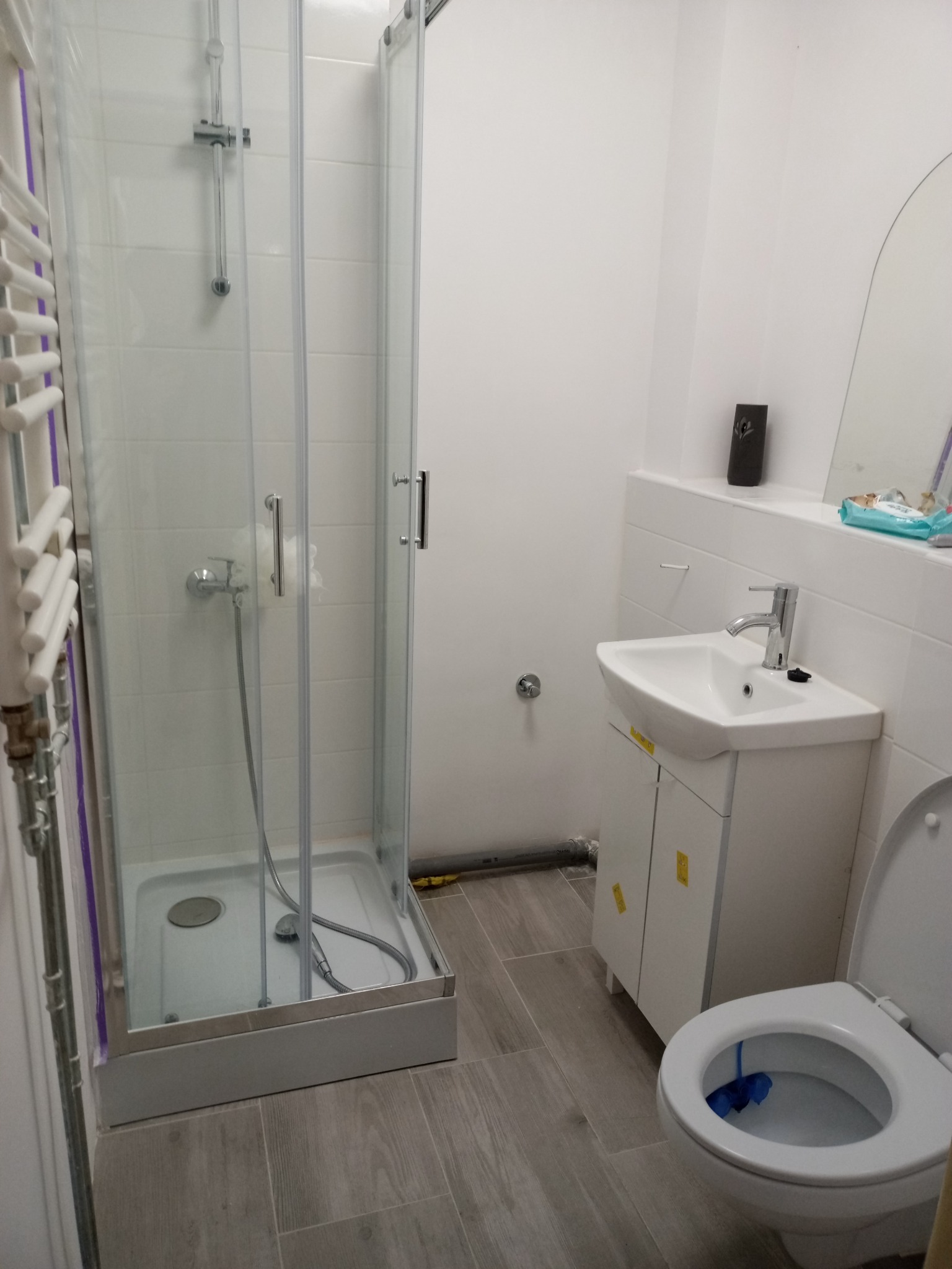 Nowoczesna łazienka z kabiną prysznicową, umywalką z szafką i toaletą, wykończona płytkami imitującymi drewno, z widoczną rurą kanalizacyjną przy podłodze.