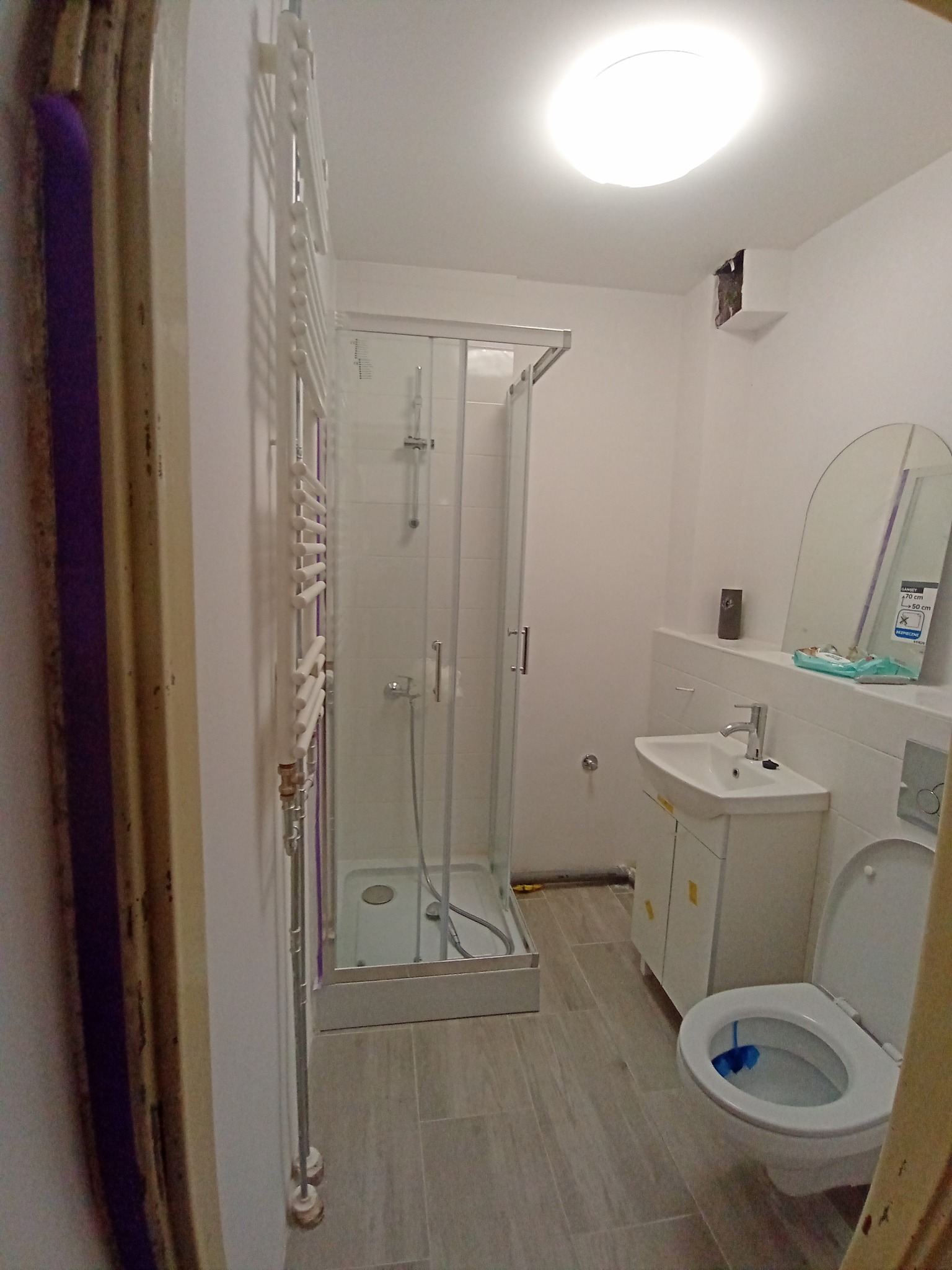 Wnętrze łazienki po remoncie: kabina prysznicowa z brodzikiem, biała umywalka z szafką, toaleta z otwartą klapą, grzejnik drabinkowy na ścianie, szare płytki podłogowe, widoczne niedoskonałości...