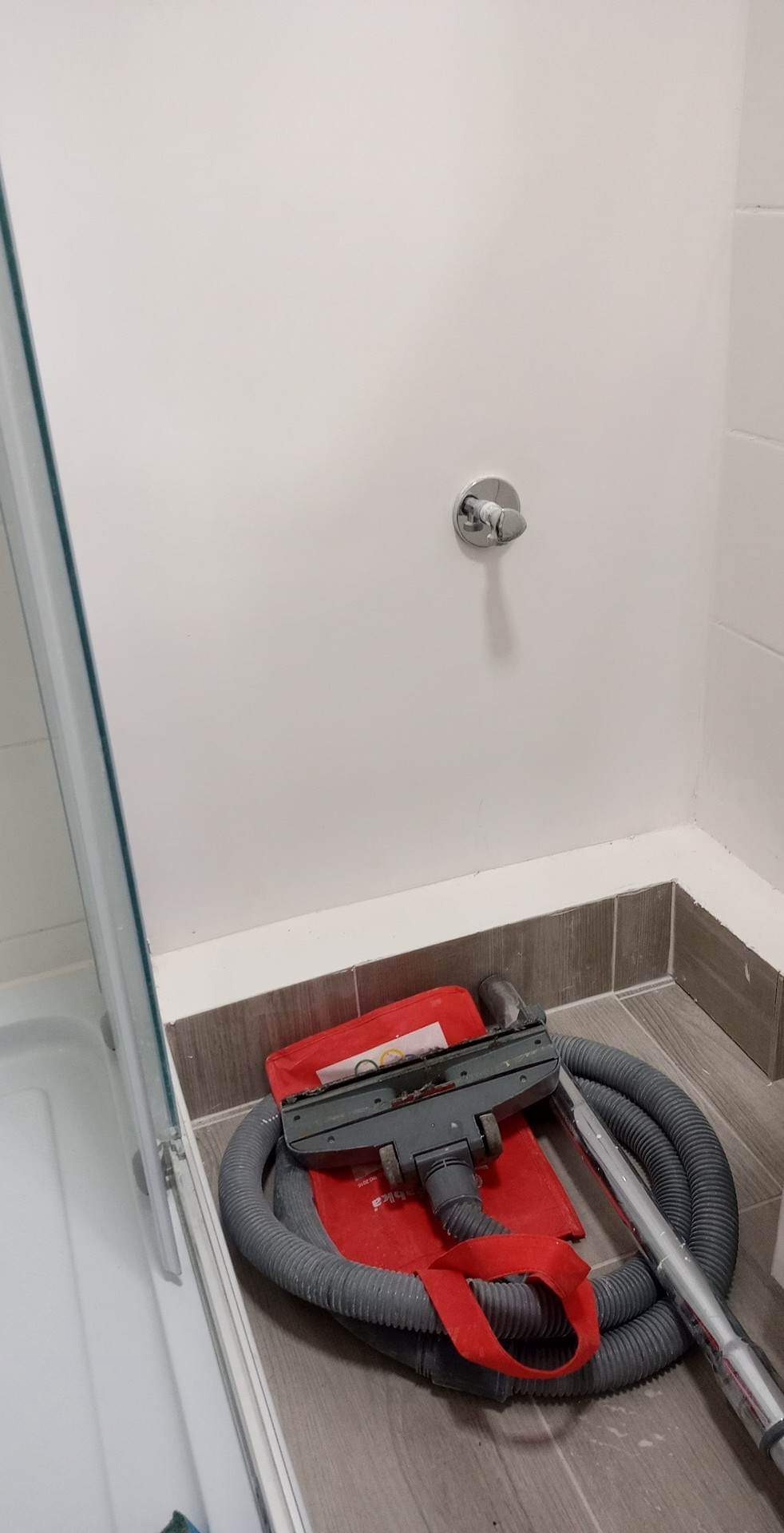 Wnętrze łazienki w trakcie sprzątania po remoncie, widoczny odkurzacz przemysłowy na podłodze obok kabiny prysznicowej, z widocznym zaworem wodnym na ścianie.