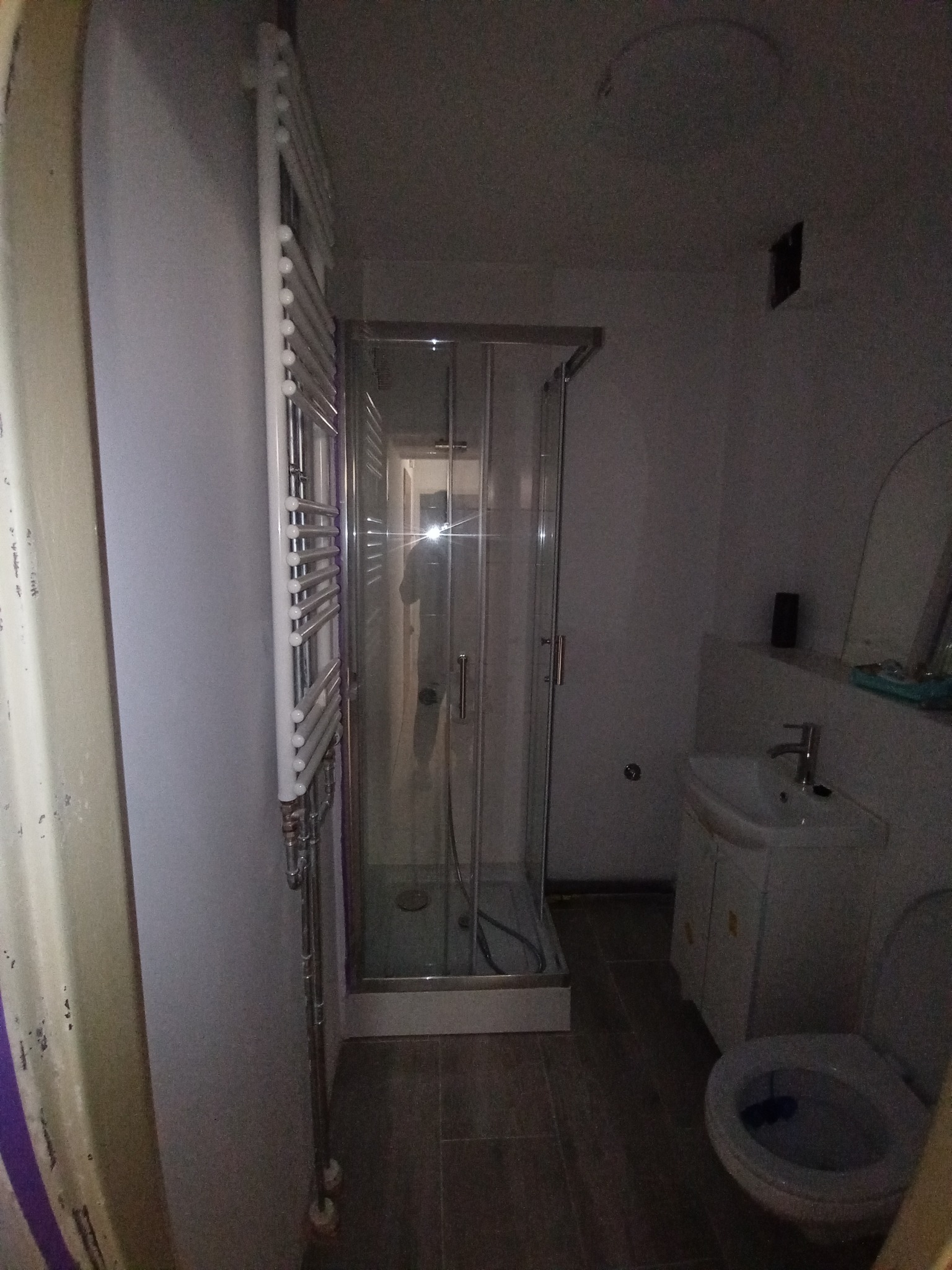 Ciemne wnętrze łazienki we Wrocławiu z kabiną prysznicową, umywalką z szafką, toaletą i grzejnikiem drabinkowym na ścianie. Widoczne ślady użytkowania i niedoskonałości wykończenia.