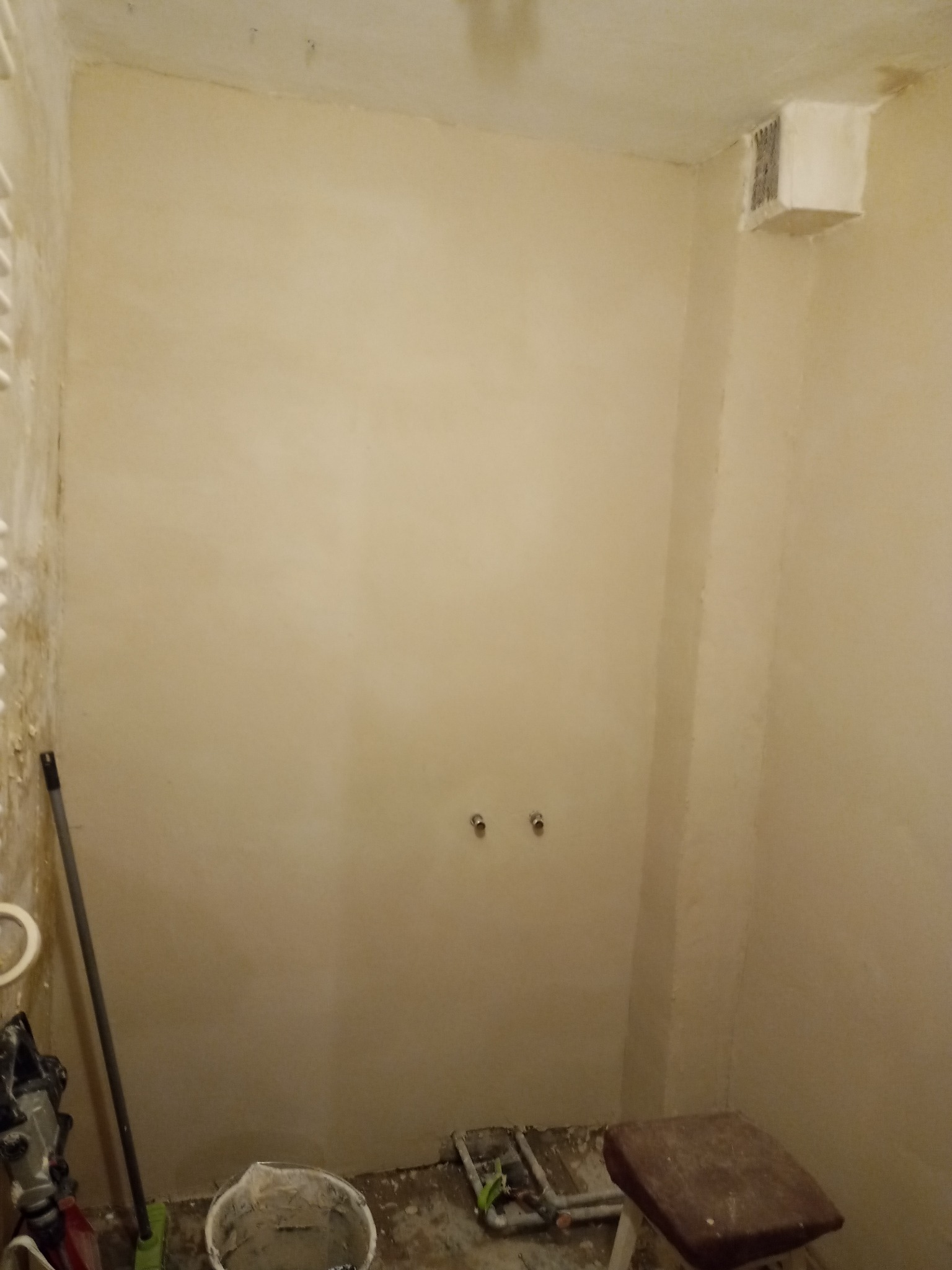 Wnętrze łazienki w trakcie prac wykończeniowych, ściany pokryte świeżo nałożoną gładzią, widoczne przyłącza wodne, wiadro z zaprawą, narzędzia i stołek, kratka wentylacyjna w rogu pomieszczenia.