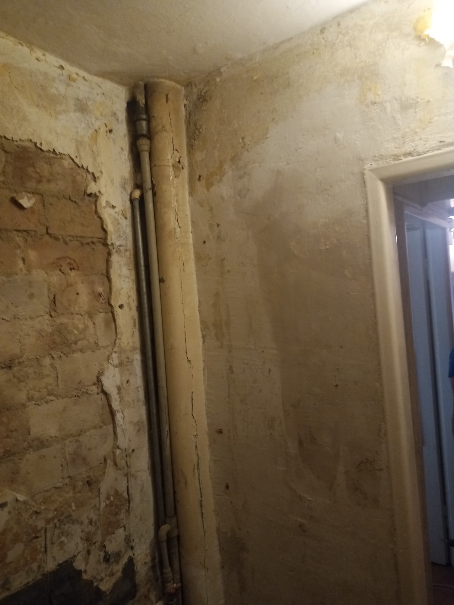 Ściana w trakcie remontu z odsłoniętą cegłą, wystającymi rurami i fragmentami starej farby, widoczne przygotowania do położenia gładzi.