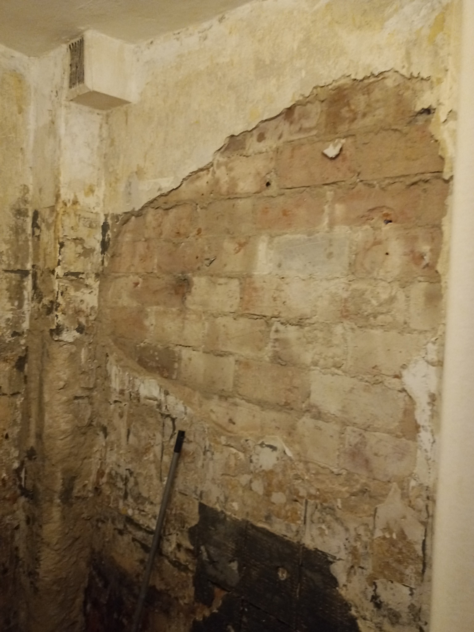 Odsłonięta ceglana ściana w trakcie skuwania tynku w pomieszczeniu, widoczny fragment wentylacji i narzędzie oparty o ścianę.