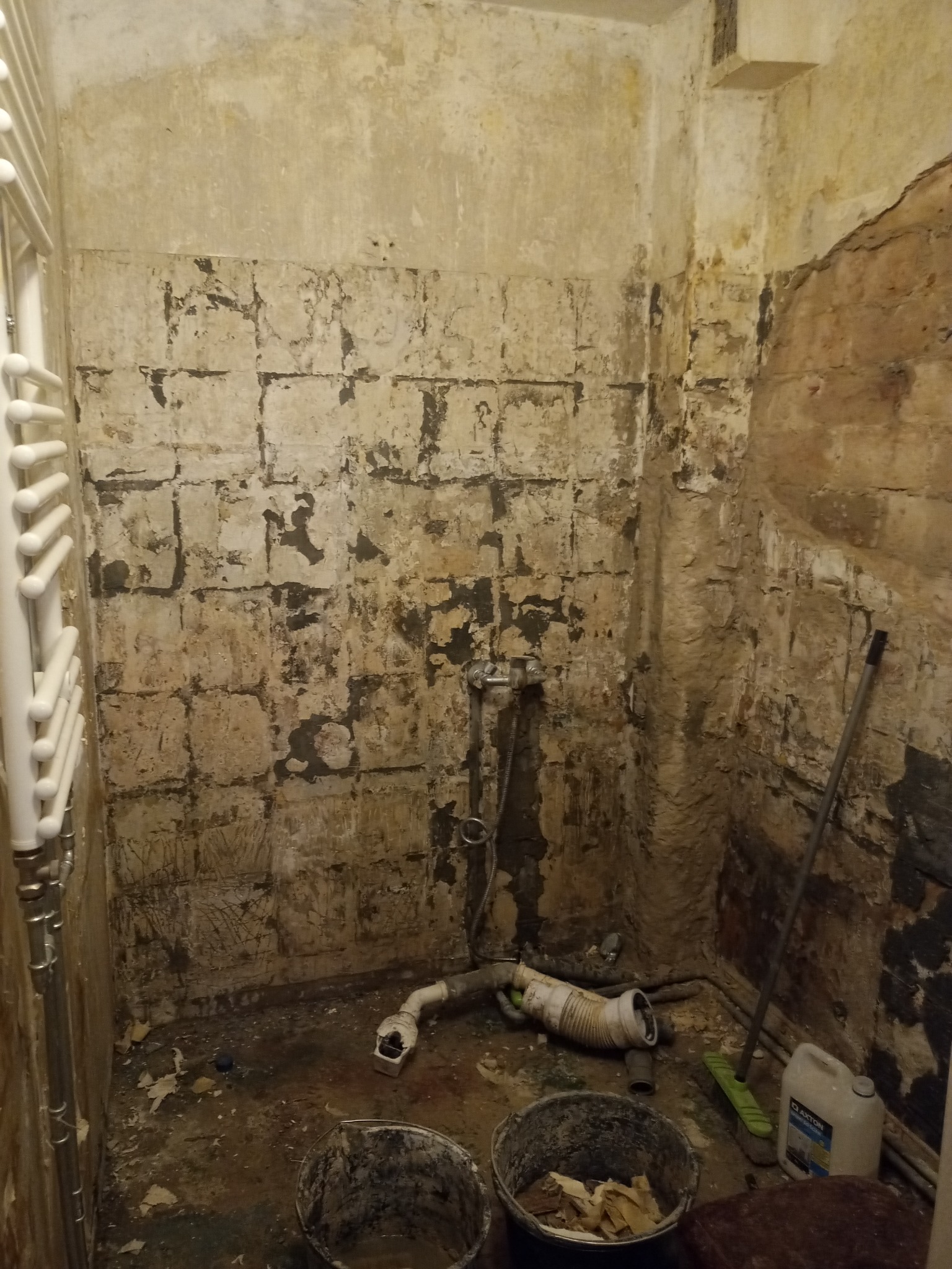 Łazienka w trakcie remontu: zerwane płytki, odsłonięte ściany, widoczne rury, wiadro z odpadami i chemia budowlana, przygotowanie do dalszych prac.