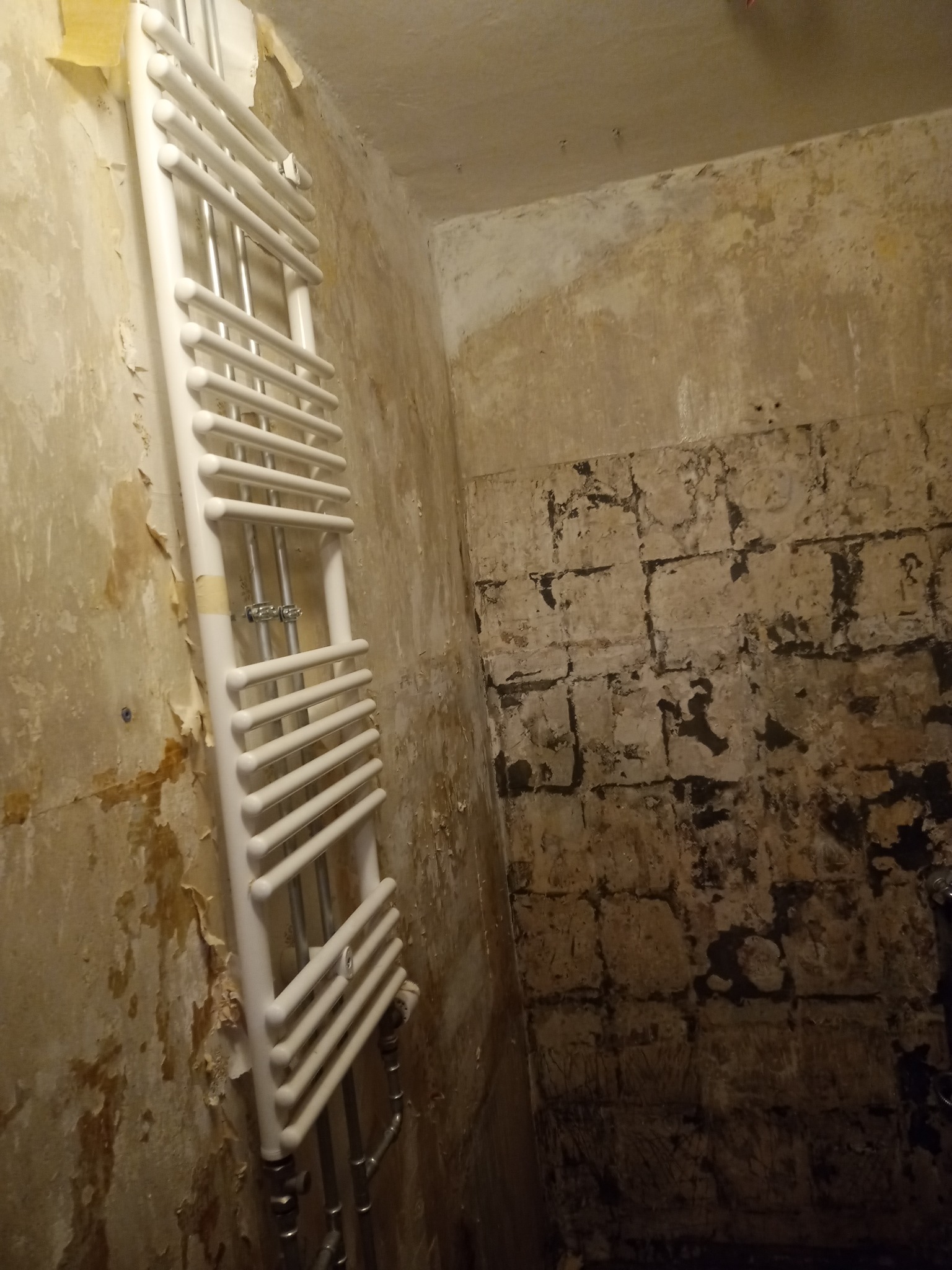 Biały grzejnik drabinkowy zamontowany na ścianie w surowej łazience w trakcie remontu, widoczne fragmenty starej farby i odsłonięta cegła.