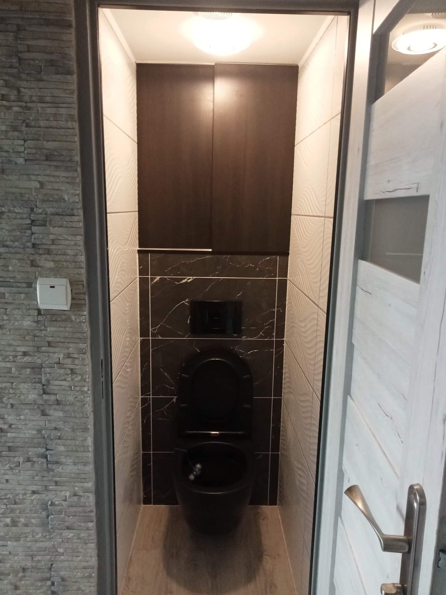 Nowoczesna, wąska toaleta z czarną armaturą i marmurowymi płytkami, oświetlona górnym światłem, widoczna zza częściowo otwartych drzwi z drewnopodobnym wykończeniem.