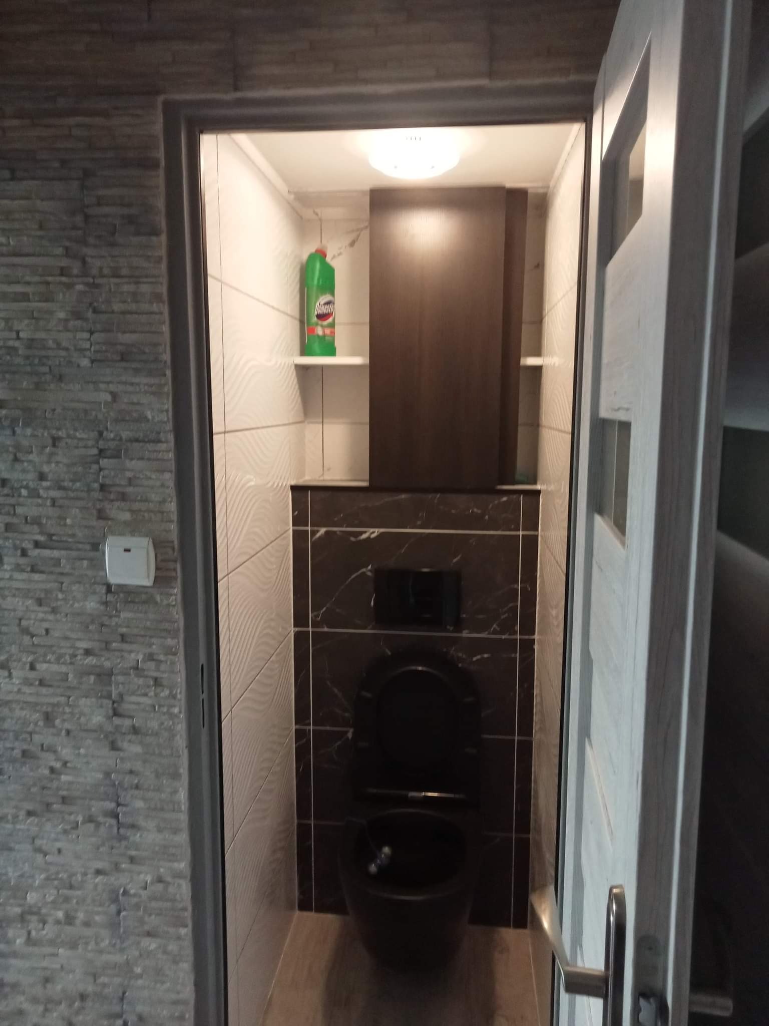 Nowoczesna łazienka z czarną miską WC, wykończona płytkami w różnych wzorach i kolorach, widoczna przez otwarte białe drzwi.