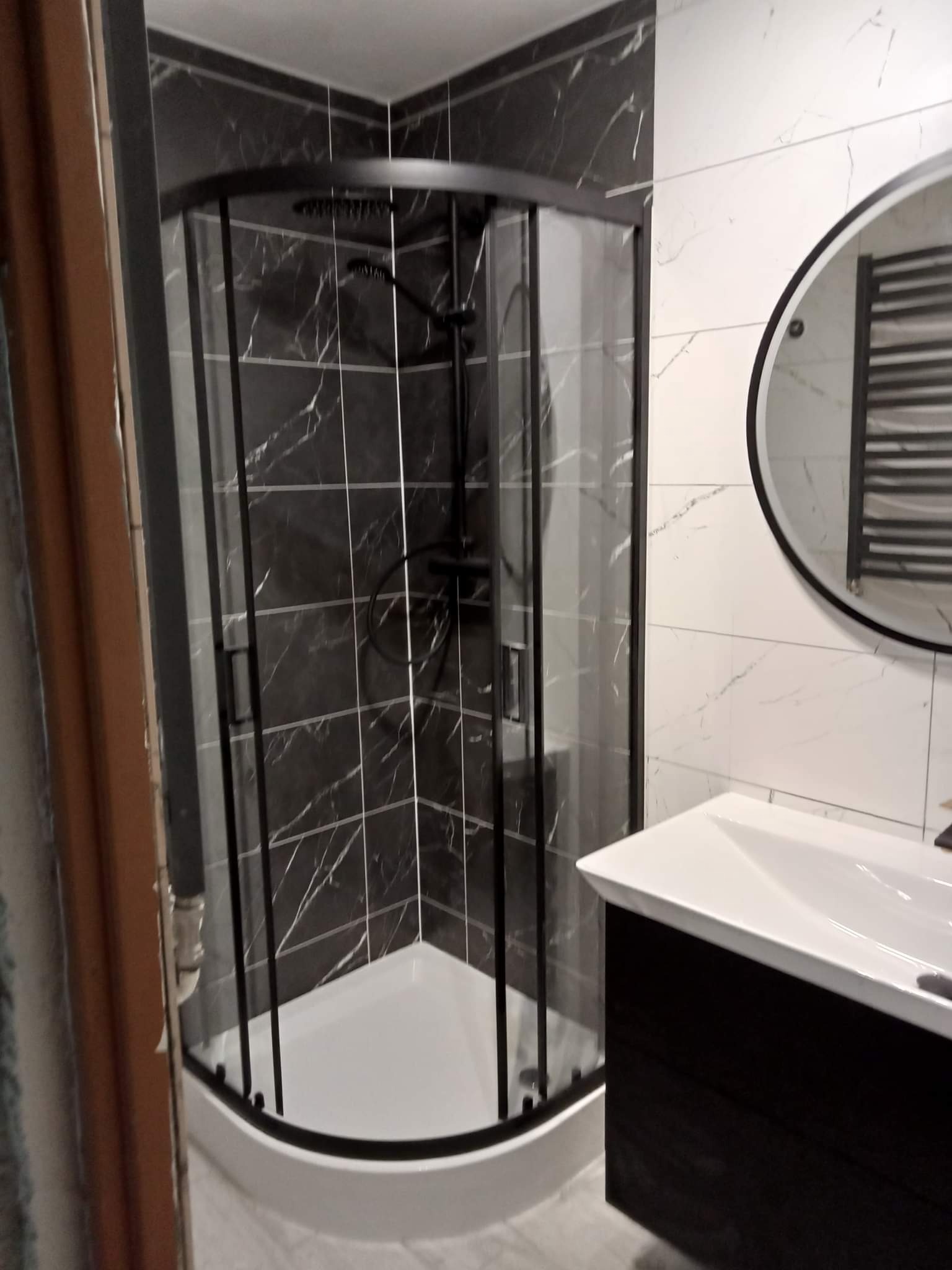 Nowoczesna łazienka z kabiną prysznicową narożną, czarną armaturą i marmurowymi płytkami w odcieniach szarości i bieli, widok na umywalkę z szafką i okrągłe lustro.