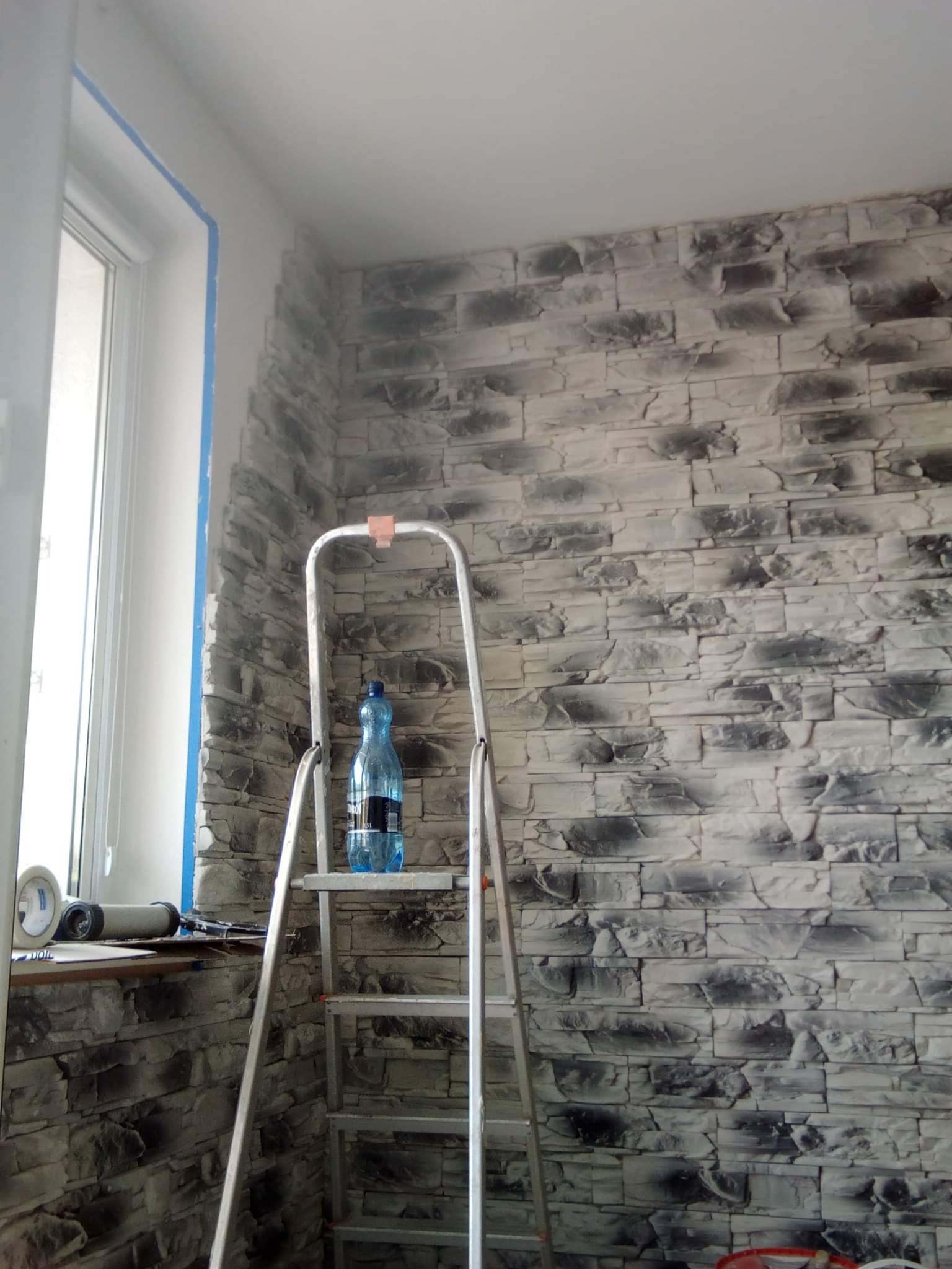 Montaż paneli ściennych imitujących kamień, widoczna drabina, butelka wody na stopniu, taśma malarska zabezpieczająca okno.