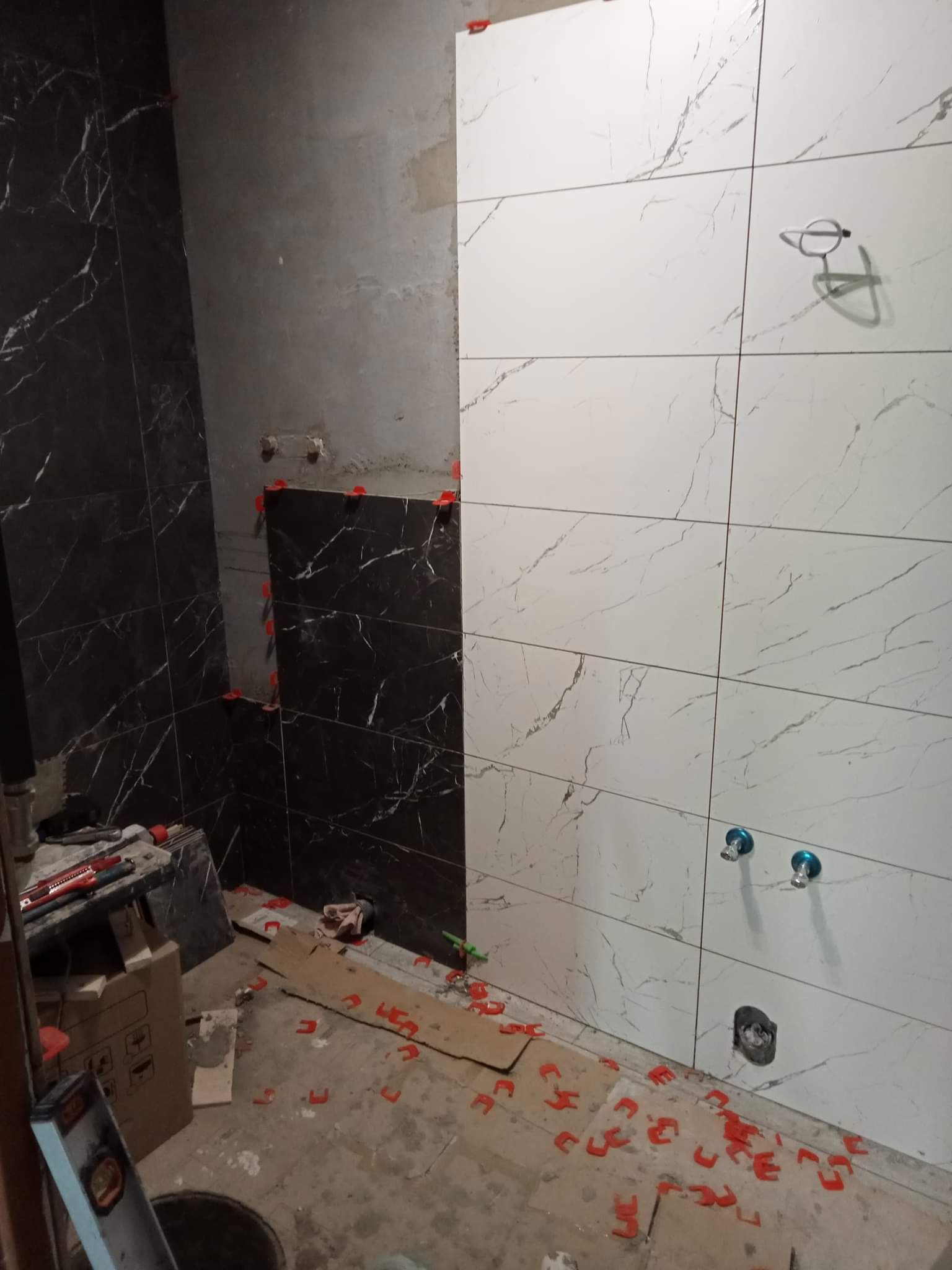 Układanie płytek imitujących marmur w łazience, widoczne krzyżyki dystansowe, przygotowane podejścia pod armaturę, fragment ściany w stanie surowym.