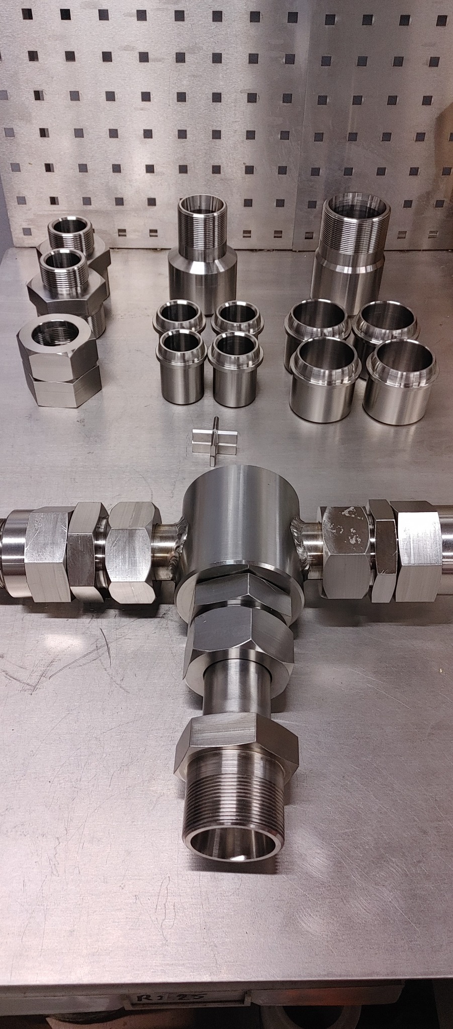 Precyzyjnie wykonane, metalowe elementy z gwintami, złączki i nakrętki o heksagonalnym kształcie, ułożone na metalowym stole roboczym z perforowaną ścianką w tle.