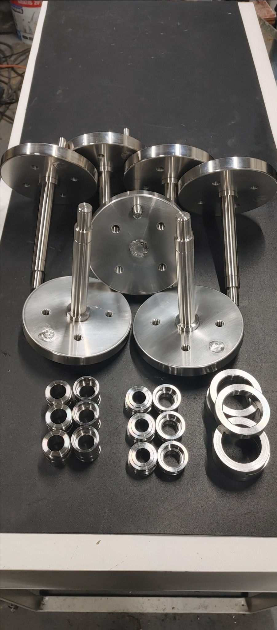 Zestaw precyzyjnie wykonanych, metalowych elementów: sześć okrągłych płyt z centralnie umieszczonym trzpieniem oraz trzy grupy pierścieni o różnych średnicach, ułożonych na ciemnym blacie.