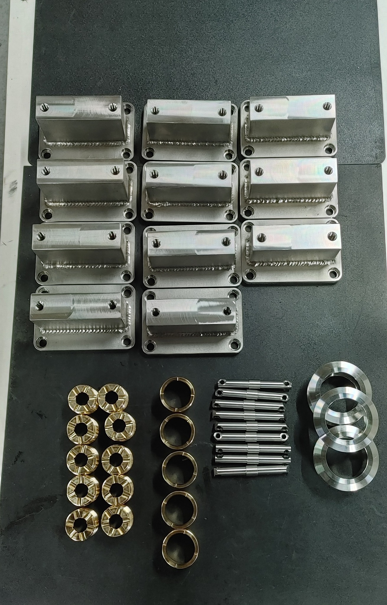 Partie obrobionych metalowych elementów: wsporniki spawane z otworami, tuleje z brązu, pręty i pierścienie na ciemnoszarym tle.
