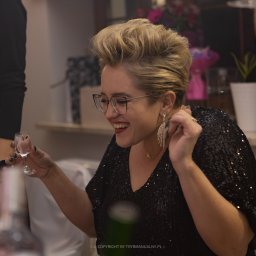 trybmanualny.pl - Uśmiechnięta kobieta w czarnej cekinowej bluzce i okularach, trzymająca kieliszek wódki podczas wydarzenia.