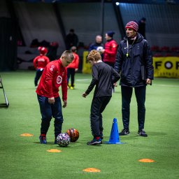 trybmanualny.pl - Trening piłkarski na hali: dwóch chłopców ćwiczy drybling między piłkami i pachołkami pod okiem trenera w czapce w paski.