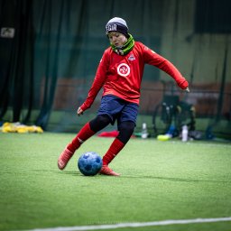 trybmanualny.pl - Młody piłkarz w czerwonej koszulce i niebieskich spodenkach kopie niebieską piłkę na zielonym boisku w hali.