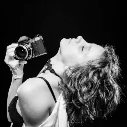 Czarno-białe zdjęcie kobiety z zamkniętymi oczami, trzymającej w rękawiczce stary aparat Praktica, z ciemnym tłem i delikatnym oświetleniem.