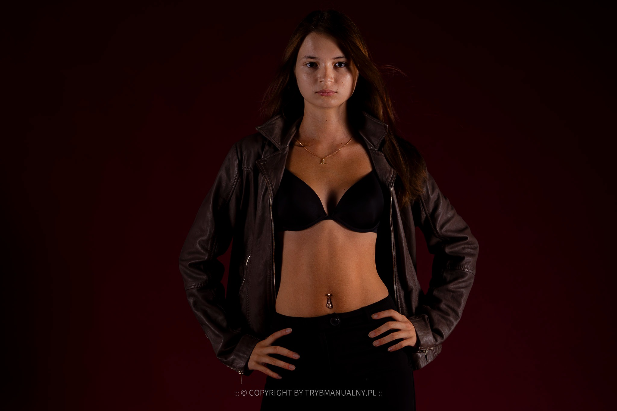 Portret młodej kobiety w czarnej bieliźnie i skórzanej kurtce, na ciemnoczerwonym tle, z rękami na biodrach i widocznym piercingiem w pępku.
