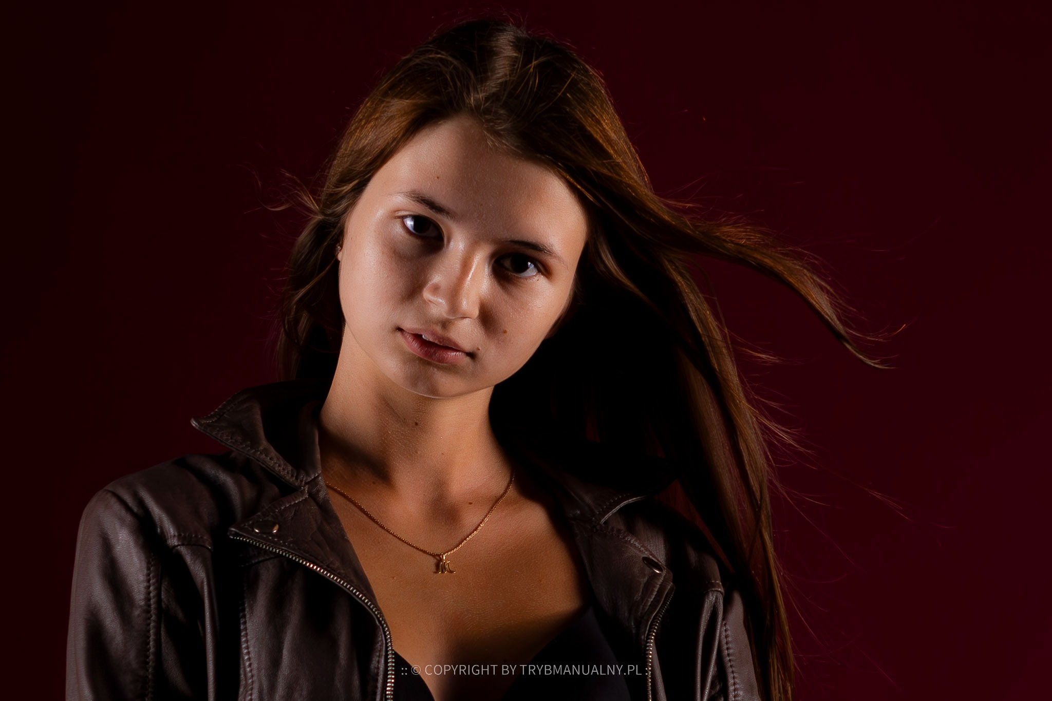 Portret młodej kobiety w skórzanej kurtce na ciemnoczerwonym tle, z włosami delikatnie rozwianymi przez wiatr, eksponujący złotą zawieszkę na szyi w kształcie litery.