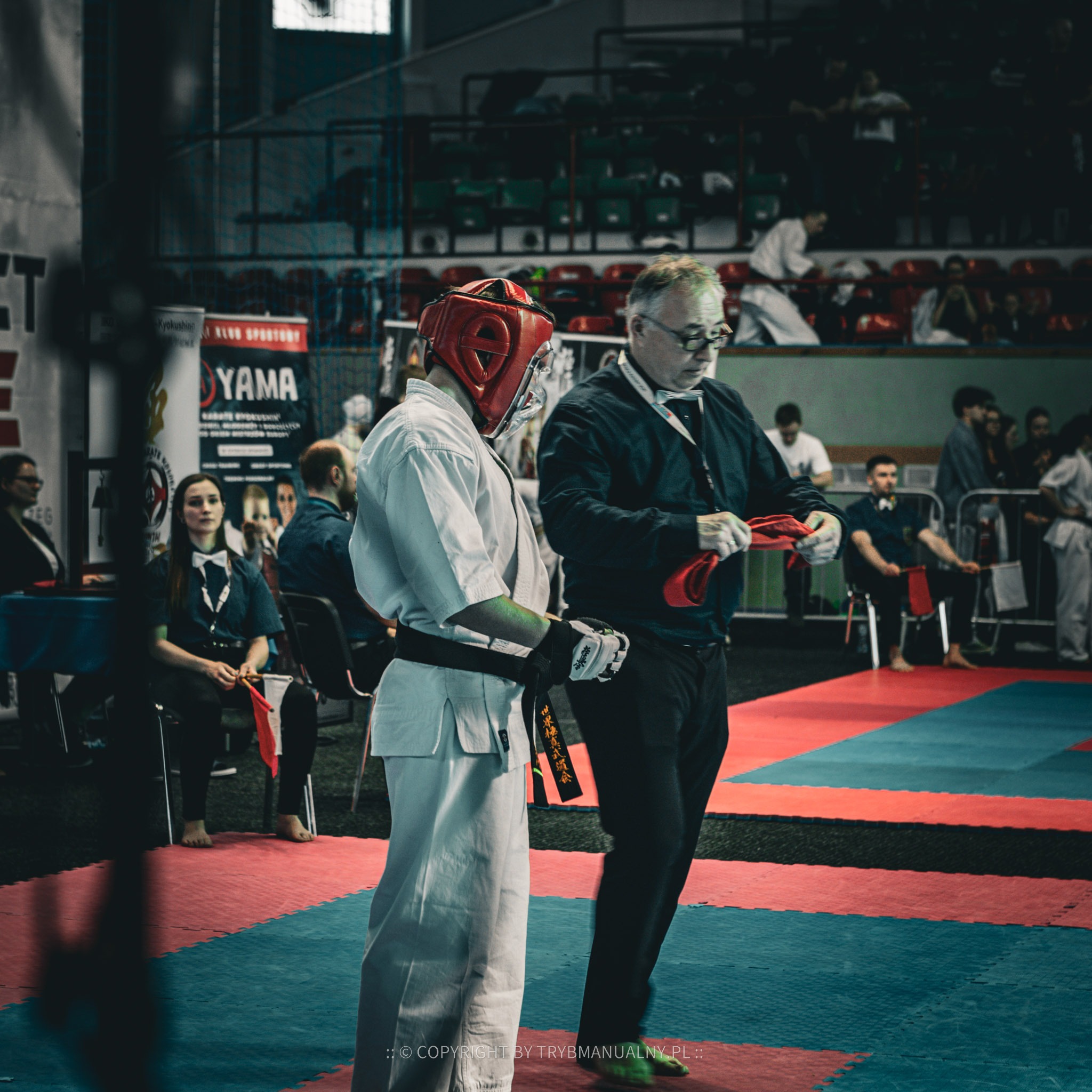 Zawodnik karate w białym kimonie i czerwonym kasku ochronnym, przygotowywany do walki przez sędziego na tle trybun z publicznością.