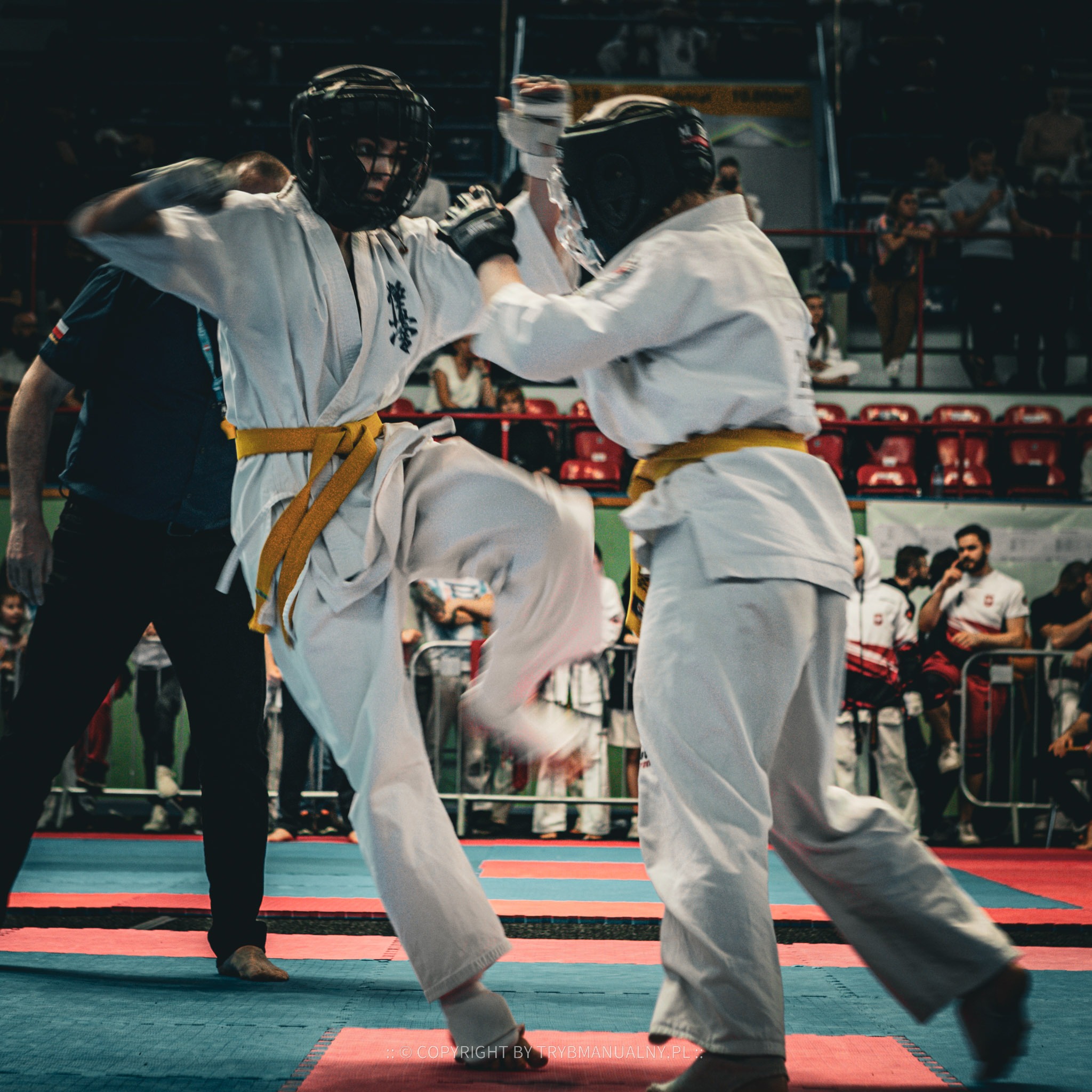 Dwóch zawodników karate w kimonach i ochraniaczach głowy podczas walki na tatami, widoczni widzowie i sędzia.