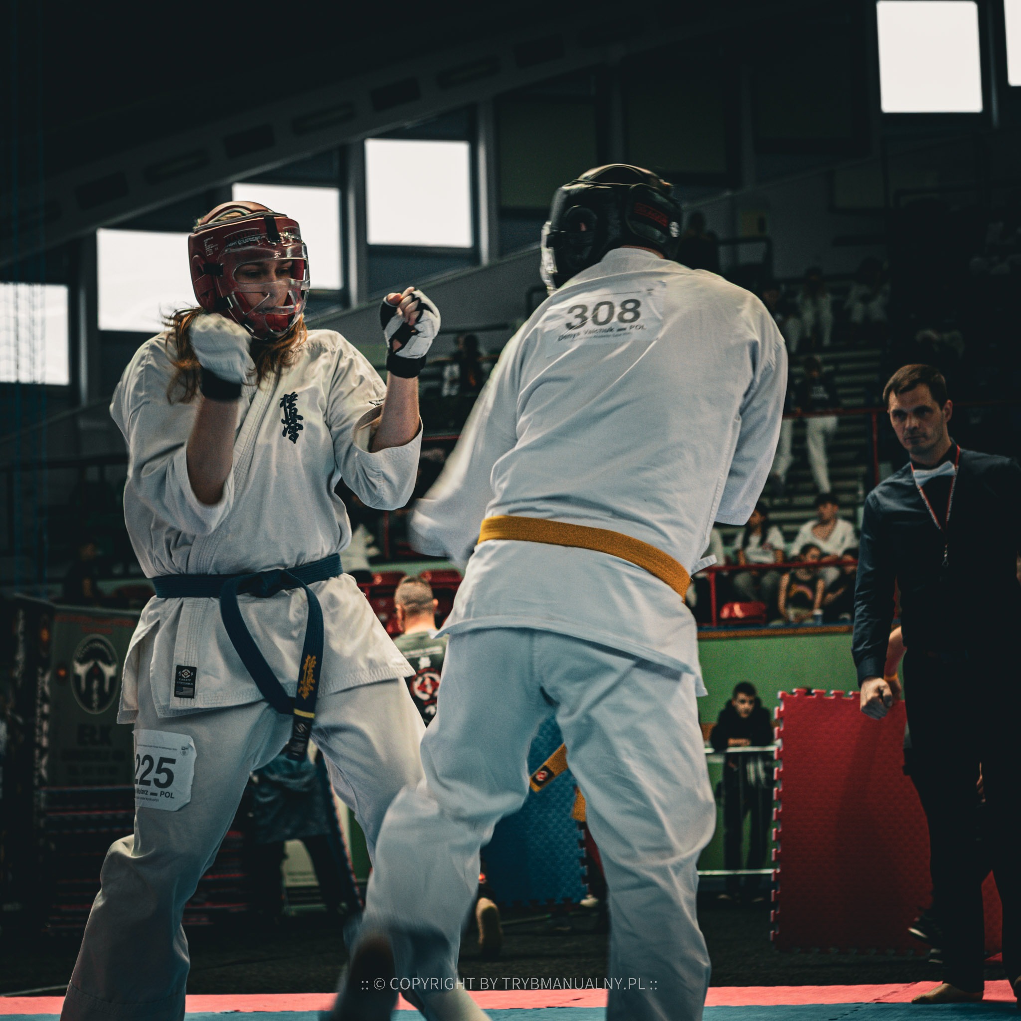 Dwóch zawodników karate w białych strojach i ochraniaczach podczas walki na zawodach, widoczny sędzia w tle.