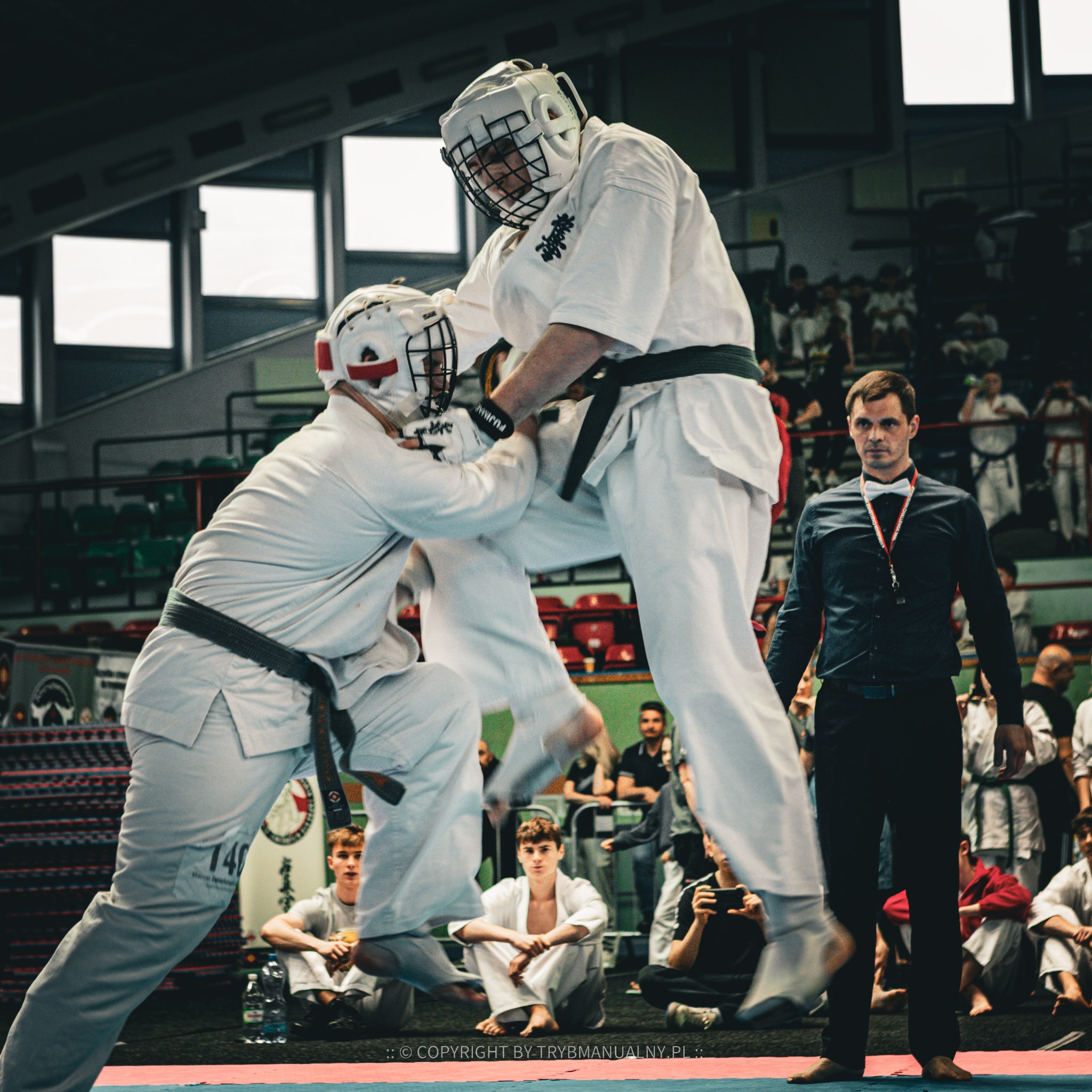 Dwóch zawodników Kyokushin Karate w białych kimonach i kaskach ochronnych podczas walki na turnieju, sędzia obserwuje, publiczność w tle.