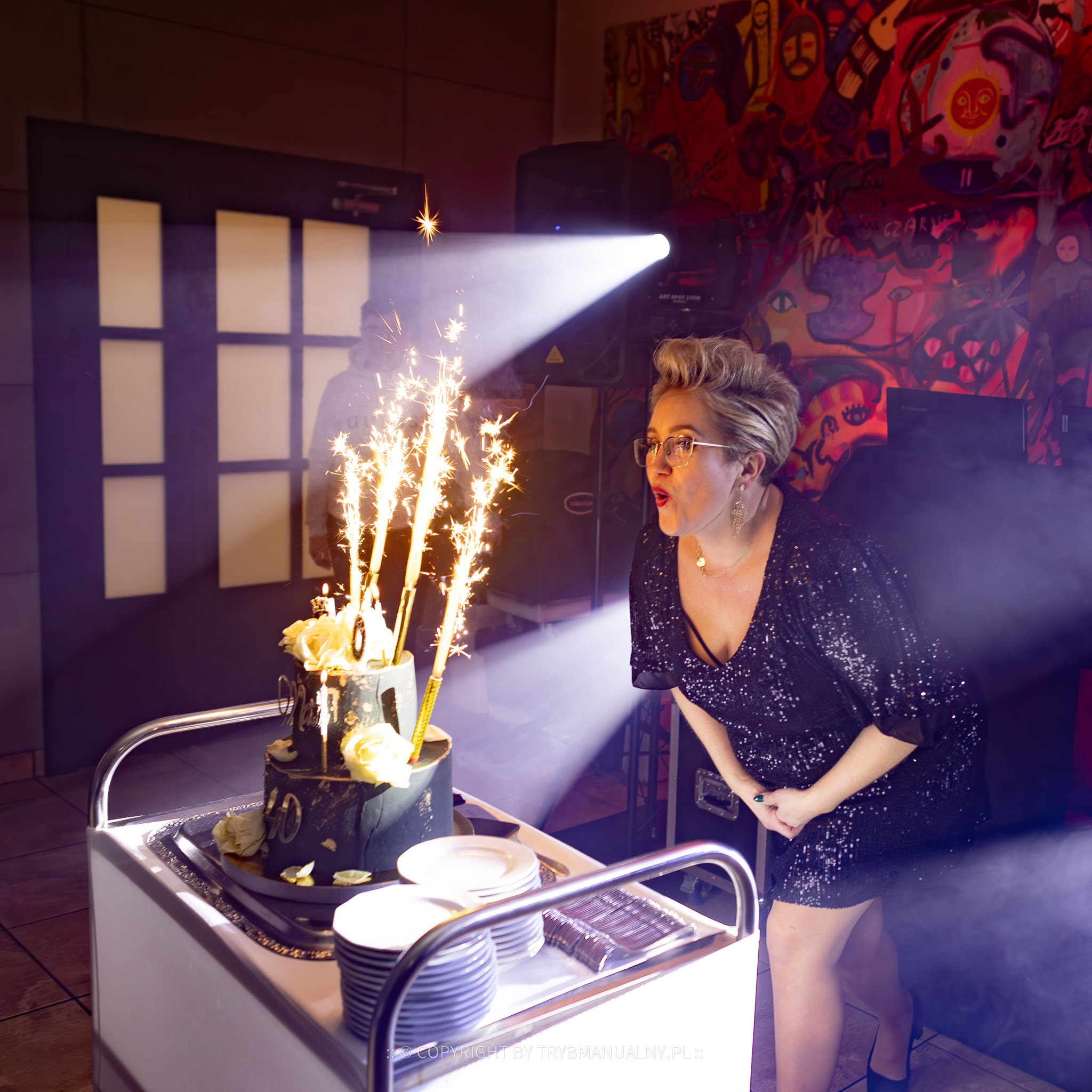 Kobieta w cekinowej sukience zdmuchuje świeczki tortowe w kształcie cyfry 40 podczas urodzinowego przyjęcia, z efektami specjalnymi dymu i świateł.