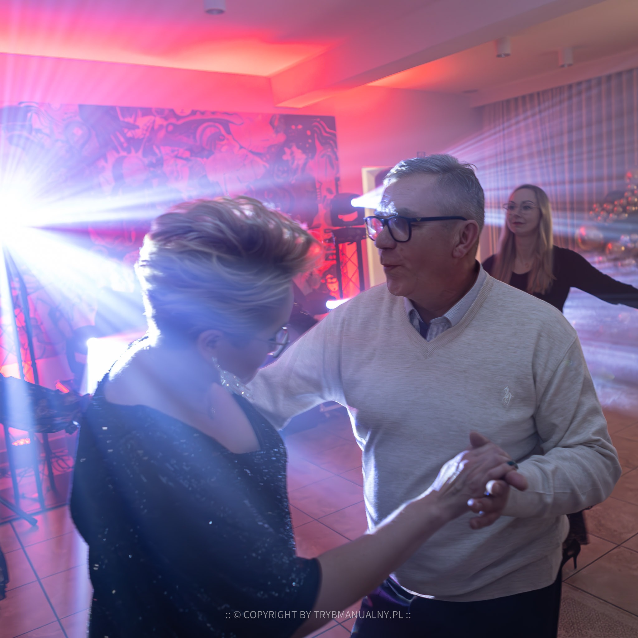 Para w średnim wieku tańczy w blasku reflektorów na sali balowej, w tle widoczna kobieta i dekoracje.