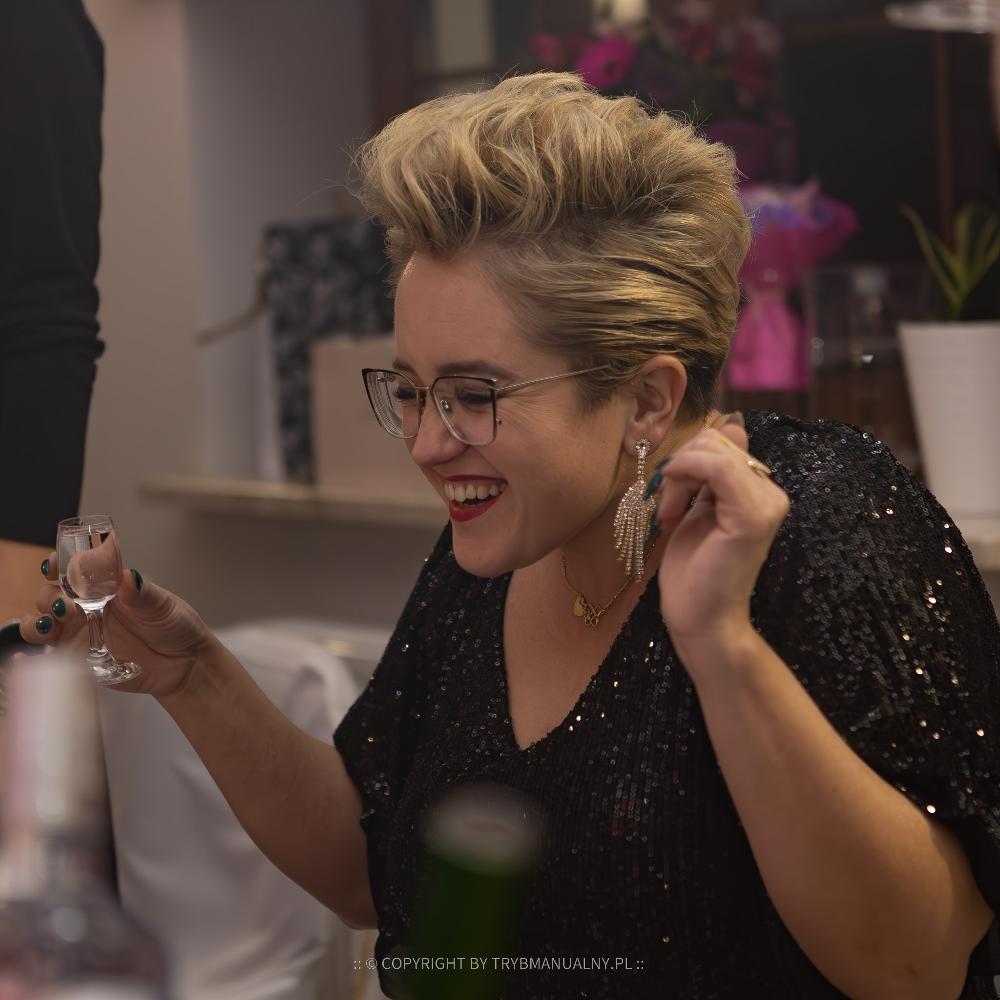 Uśmiechnięta kobieta w czarnej cekinowej bluzce i okularach, trzymająca kieliszek wódki podczas wydarzenia.