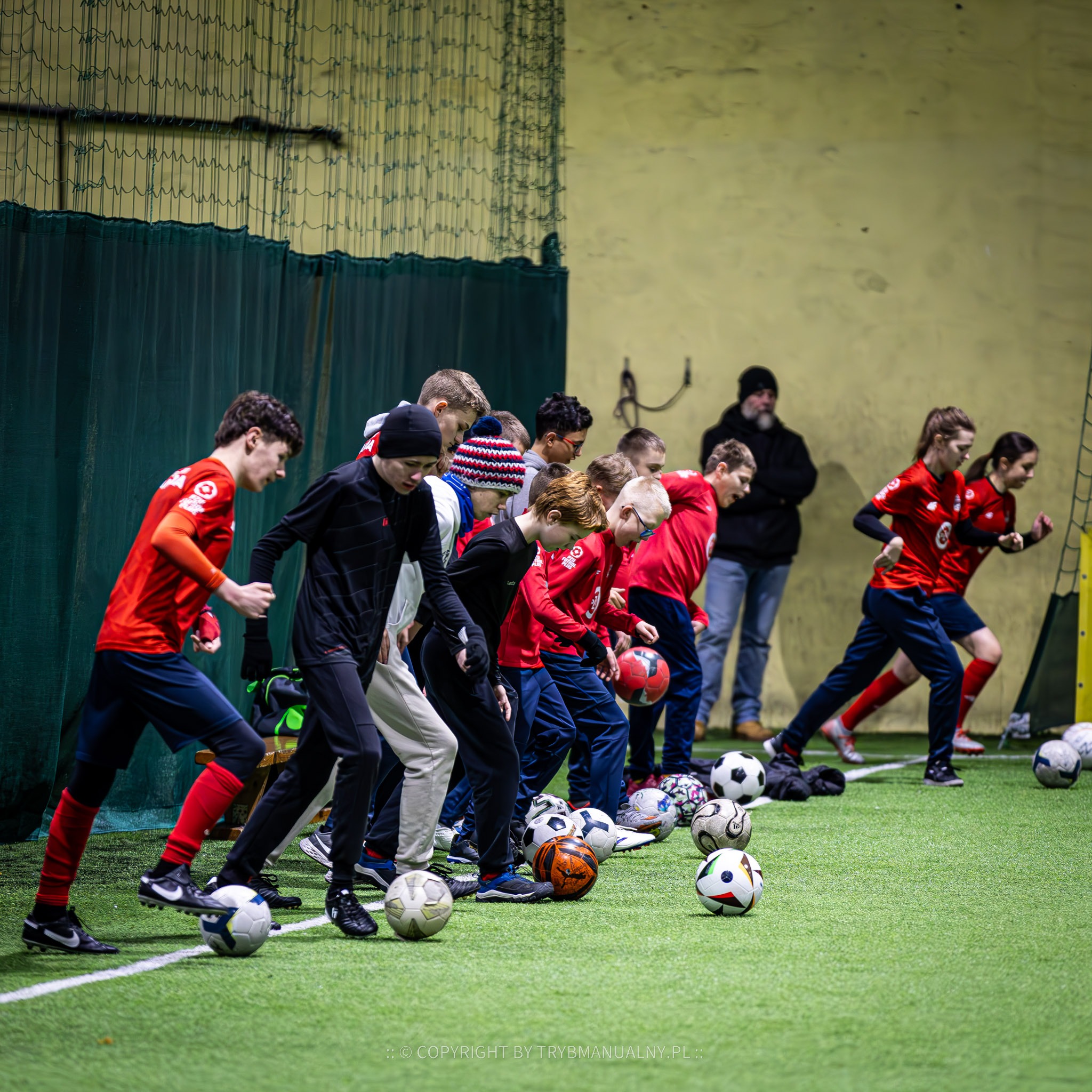 Grupa nastolatków w czerwonych koszulkach i sportowych spodniach w trakcie treningu piłkarskiego na hali, z piłkami u stóp, pod nadzorem trenera w tle.