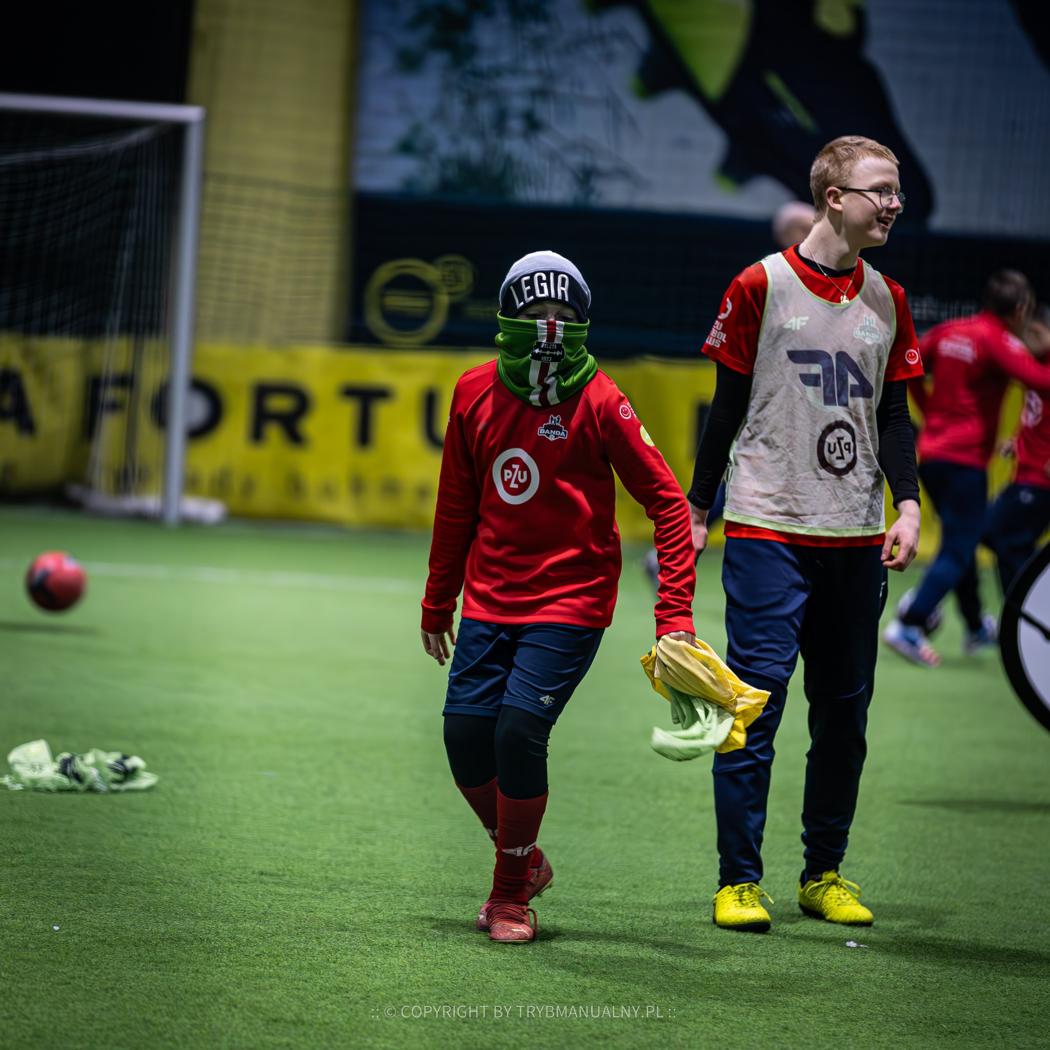 Młodzi piłkarze w strojach sportowych na hali, jeden z czapką i kominem Legii Warszawa, drugi z uśmiechem i w okularach.