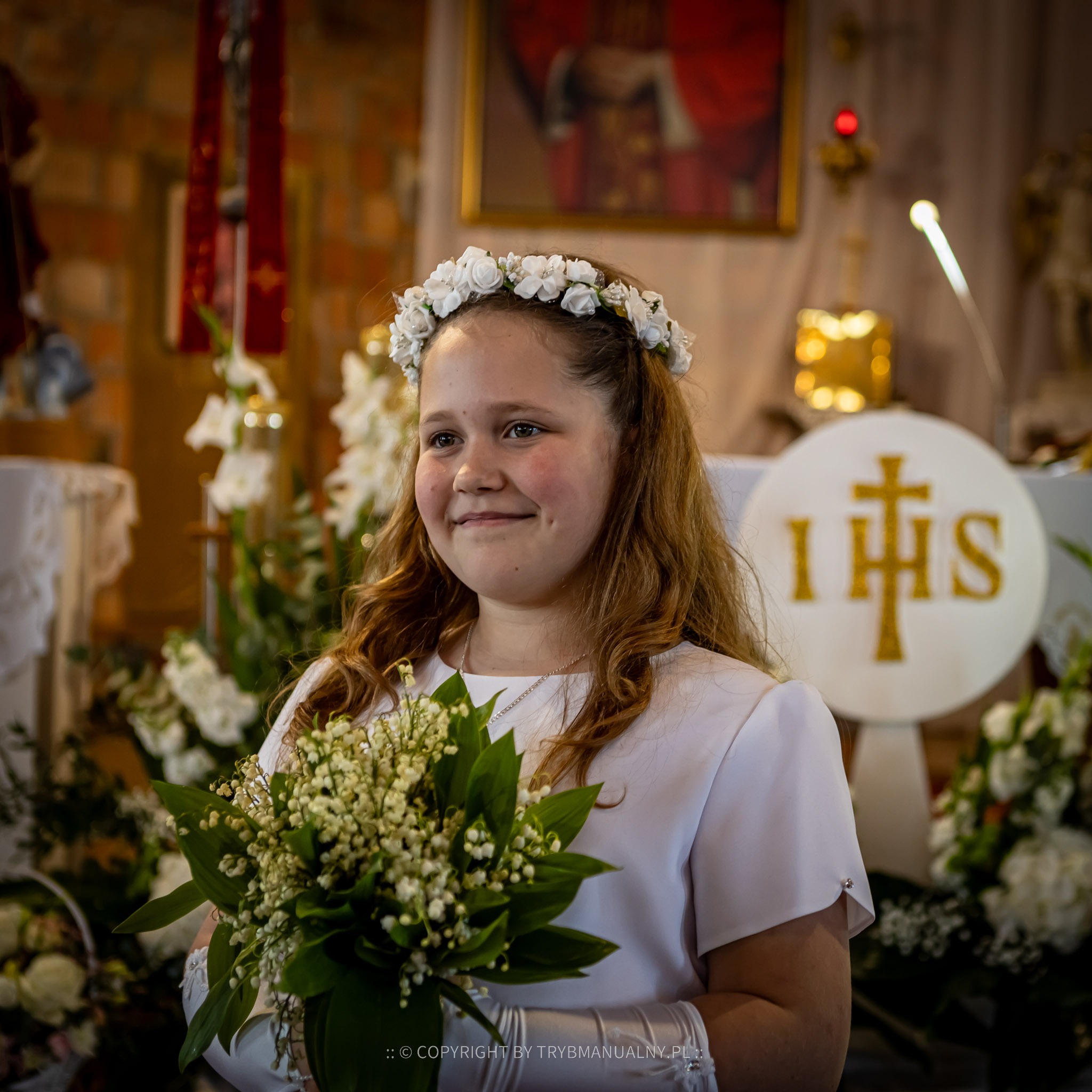 Uśmiechnięta dziewczynka w wianku z białych kwiatów, trzymająca bukiet konwalii w kościele podczas uroczystości Pierwszej Komunii Świętej, na tle ołtarza.